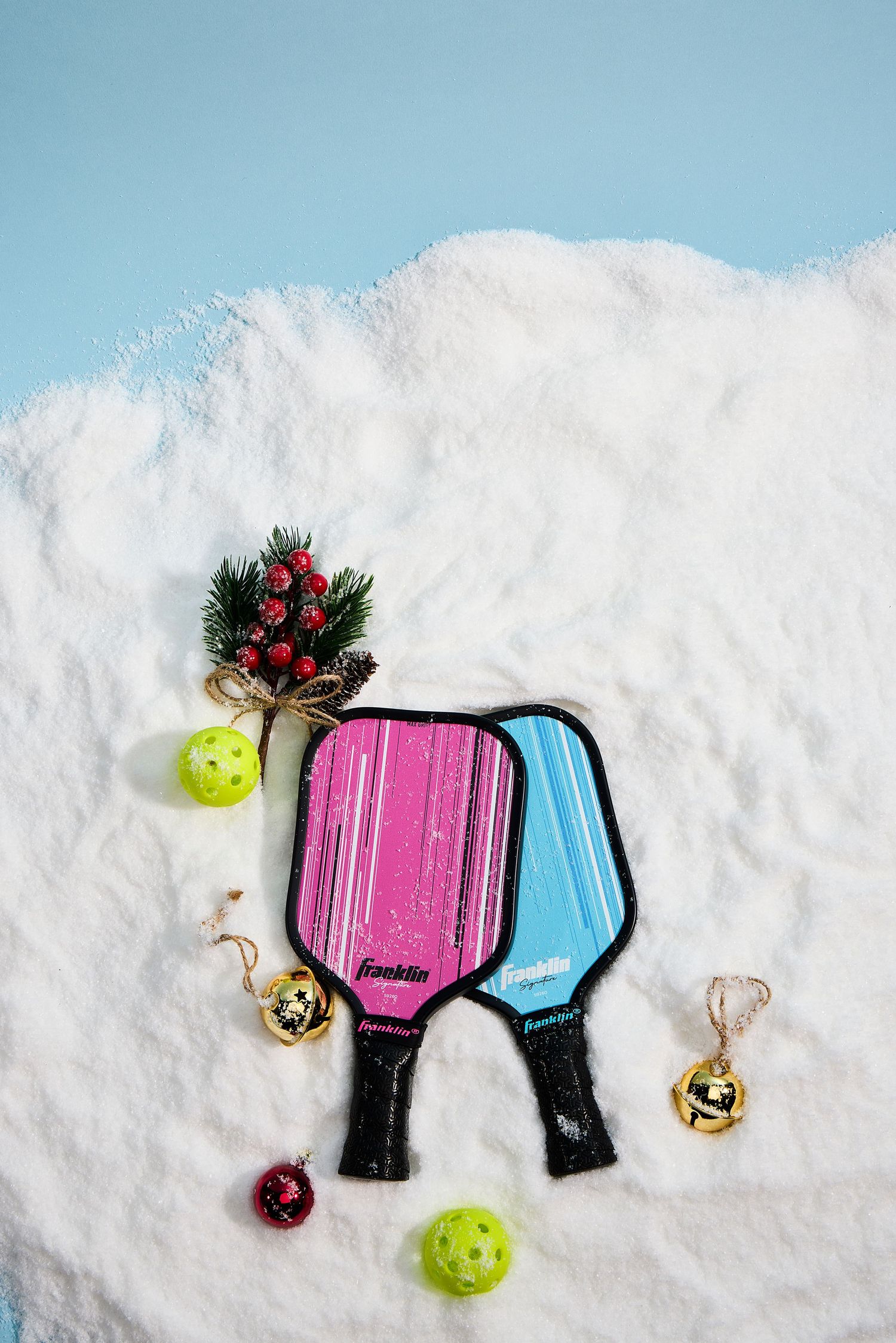 Pink and blue pickleball paddles arranged on snow with holiday ornaments and pine decorations against a light blue sky.