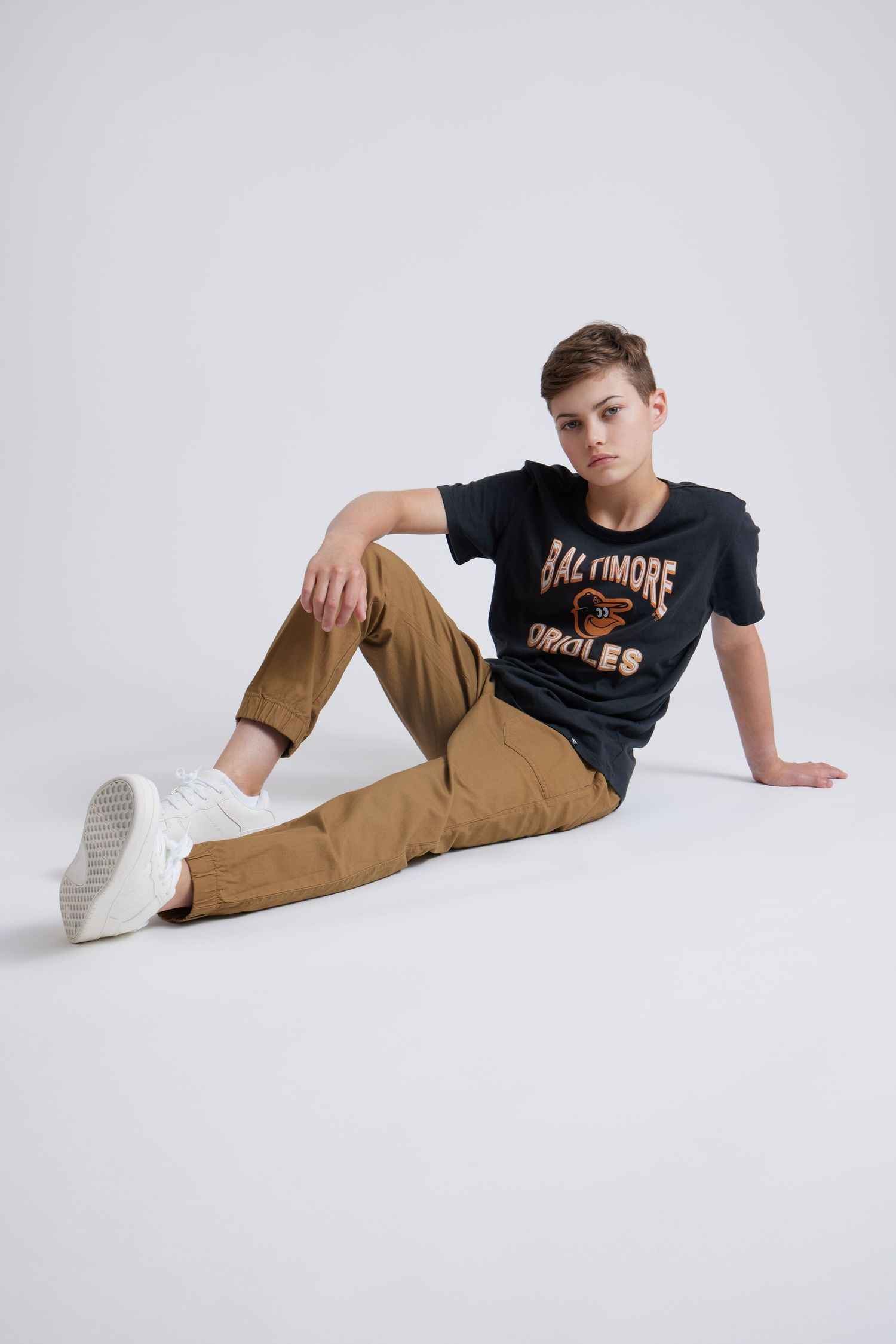 Model in black t-shirt and khaki pants poses against white backdrop in fashion photoshoot series.