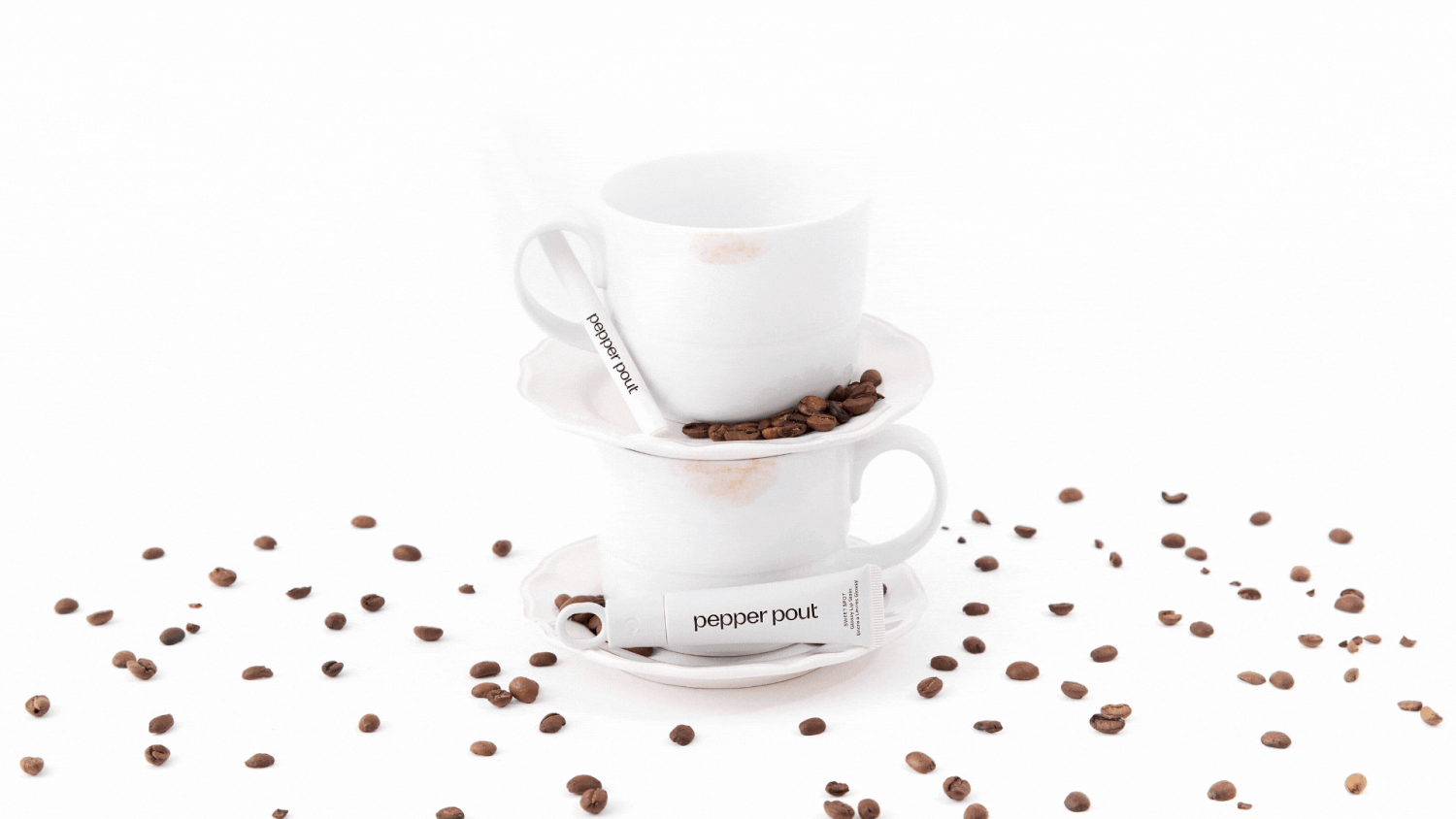 Two white coffee cups stacked on a saucer with scattered roasted coffee beans on a white background.