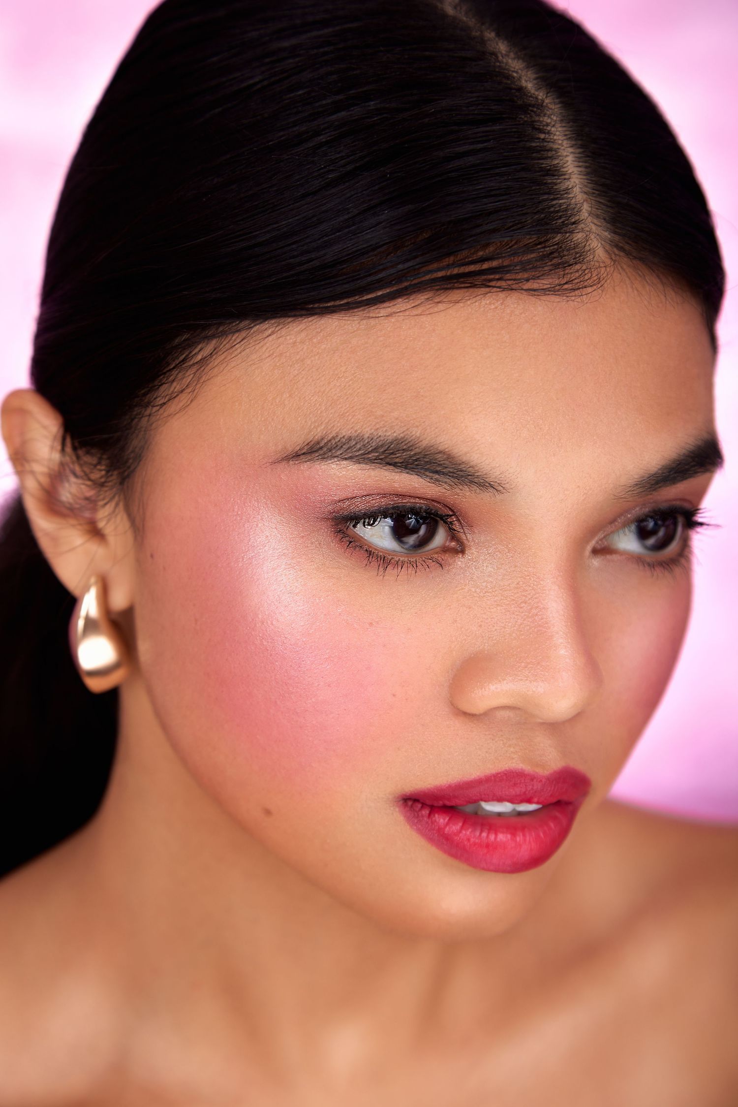 Close-up beauty portrait with bright red lipstick, pearl earrings and glowing makeup against pink background.