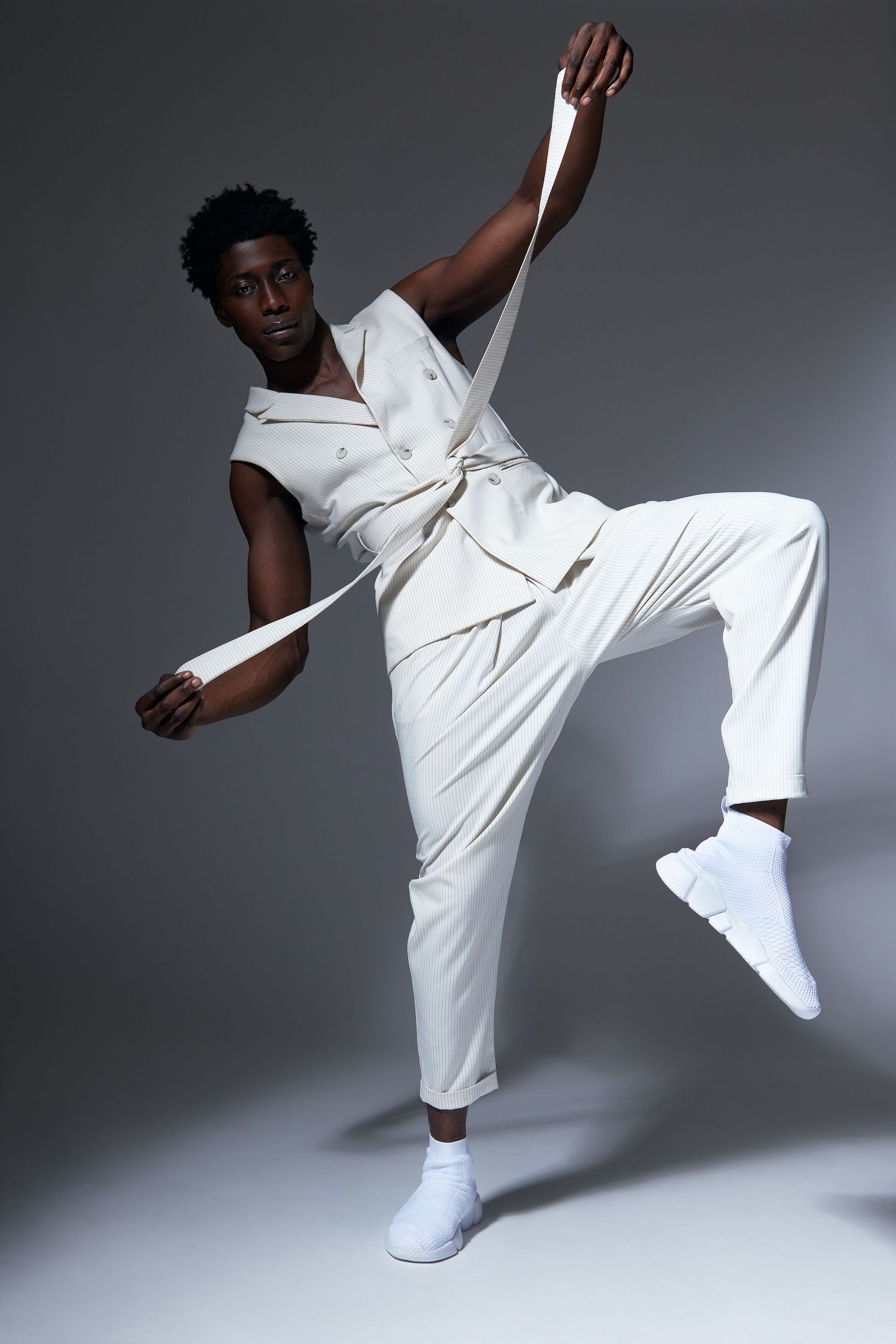 Man in white sleeveless suit performs dynamic movements against a gray studio backdrop.