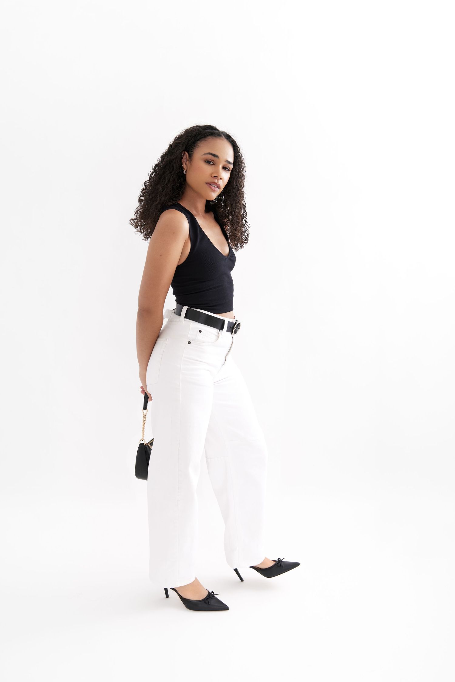 A person in a black sleeveless top and white pants poses against a white background with heels and a small purse.