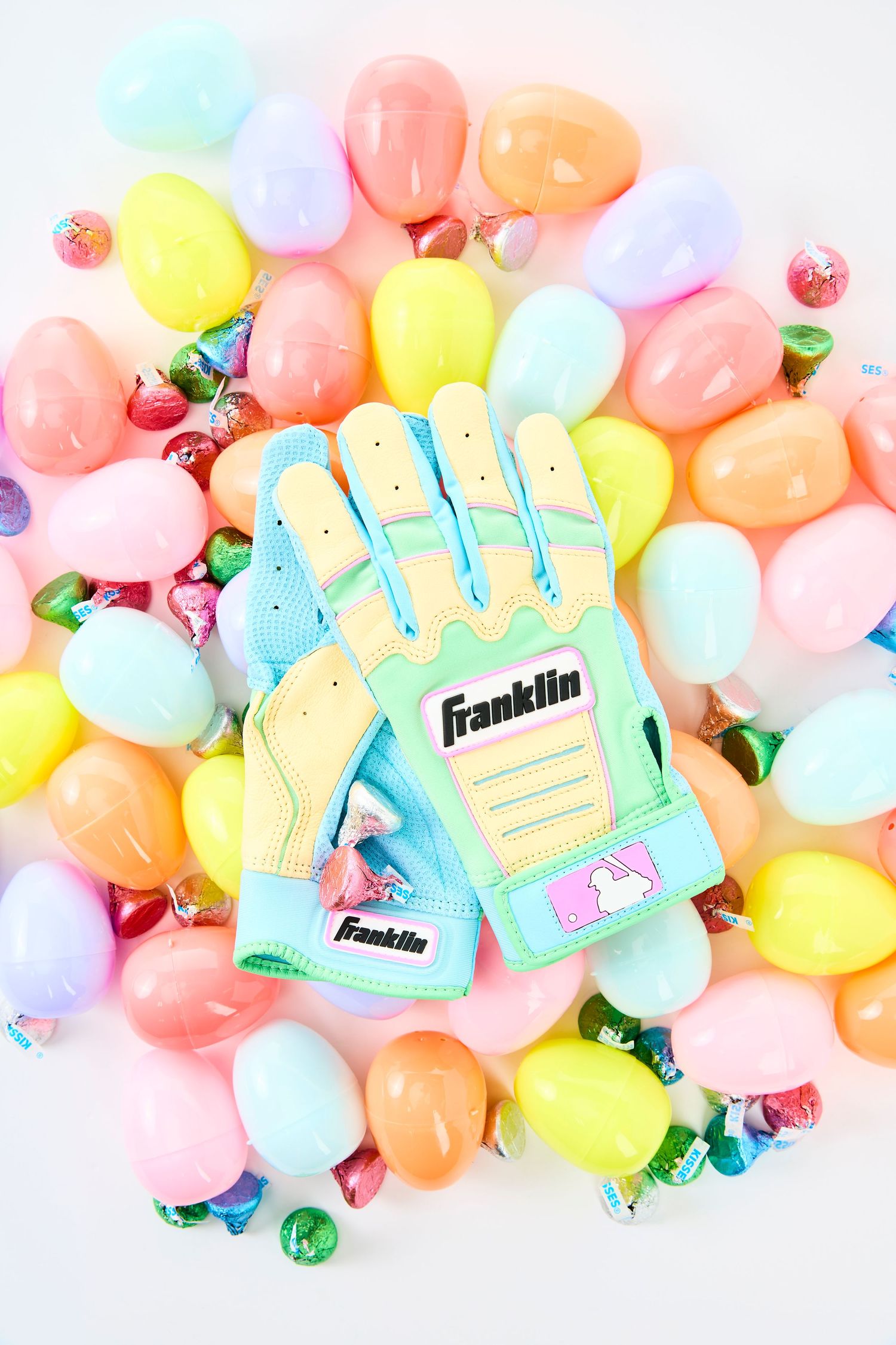 Colorful plastic Easter eggs scattered with green Franklin baseball gloves against a soft pink background.