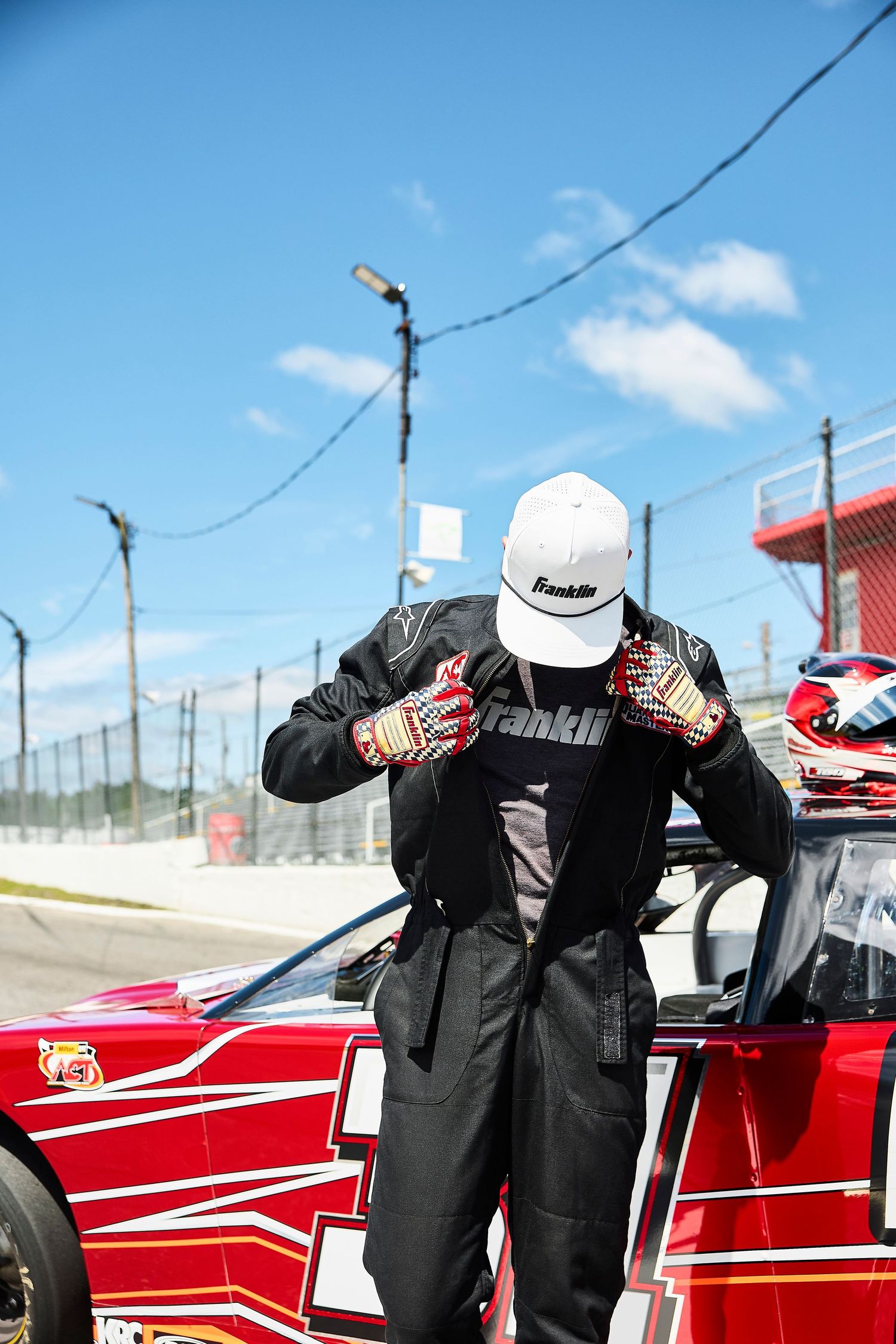 Racecar driving wearing Franklin Sports Speedway gloves and white Franklin hat in front of a racecar on a racetrack.