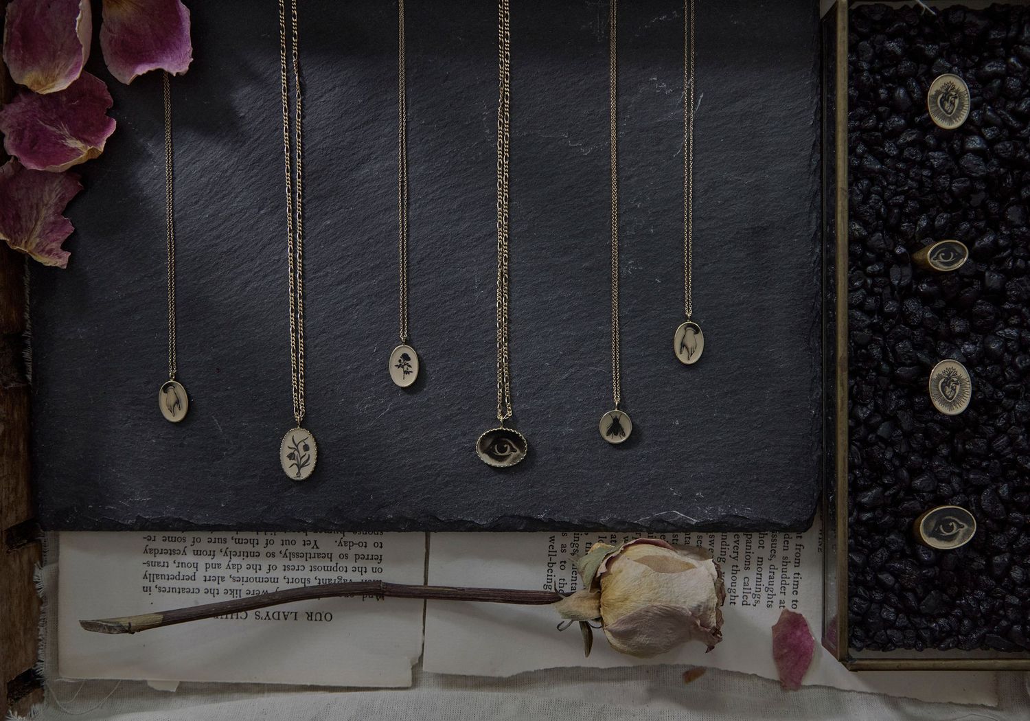 Gold oval pendant necklaces with evil eye and floral designs displayed on slate board with dried rose and book.