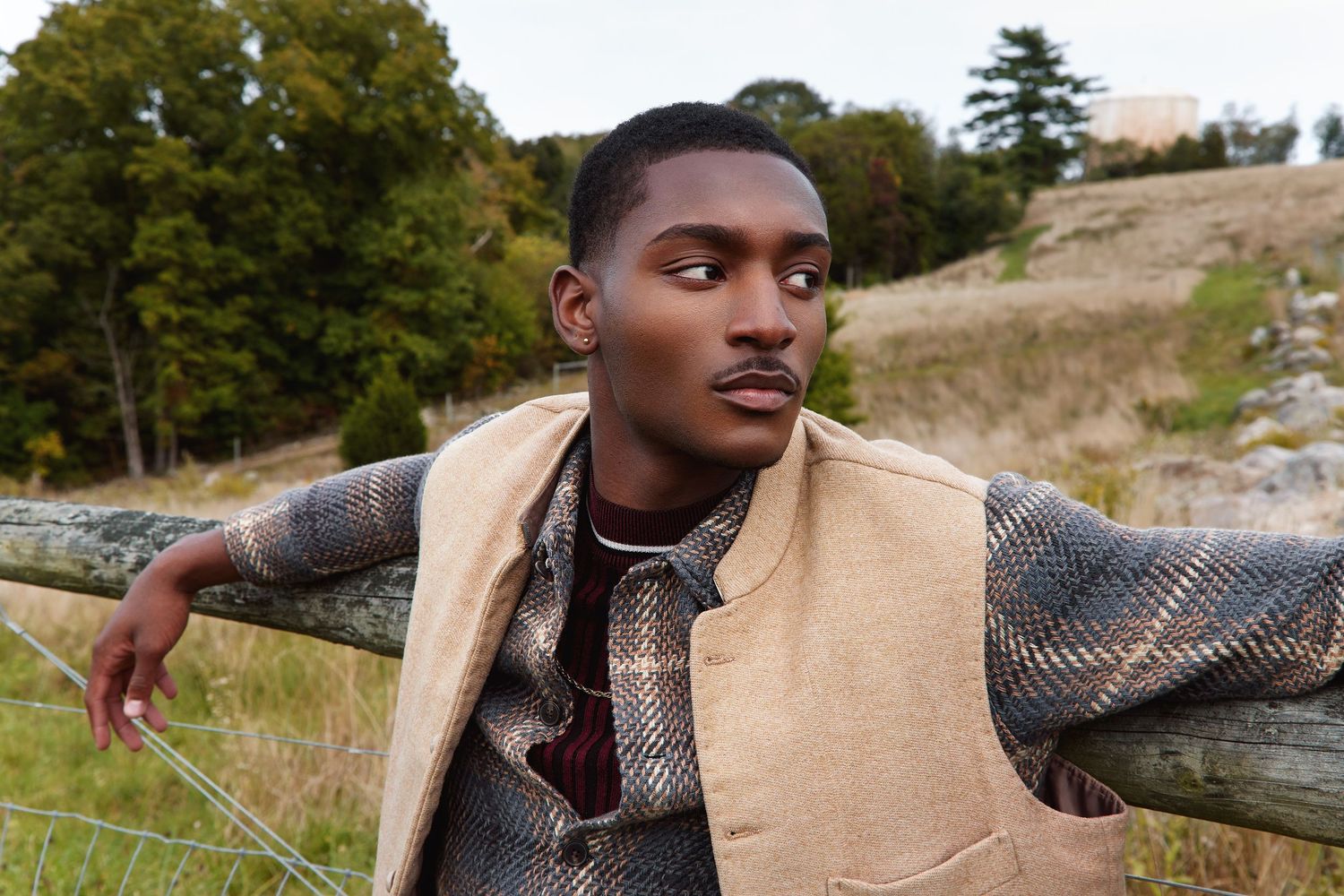 A person wearing a brown jacket poses outdoors in a natural setting with trees and hills in the background.