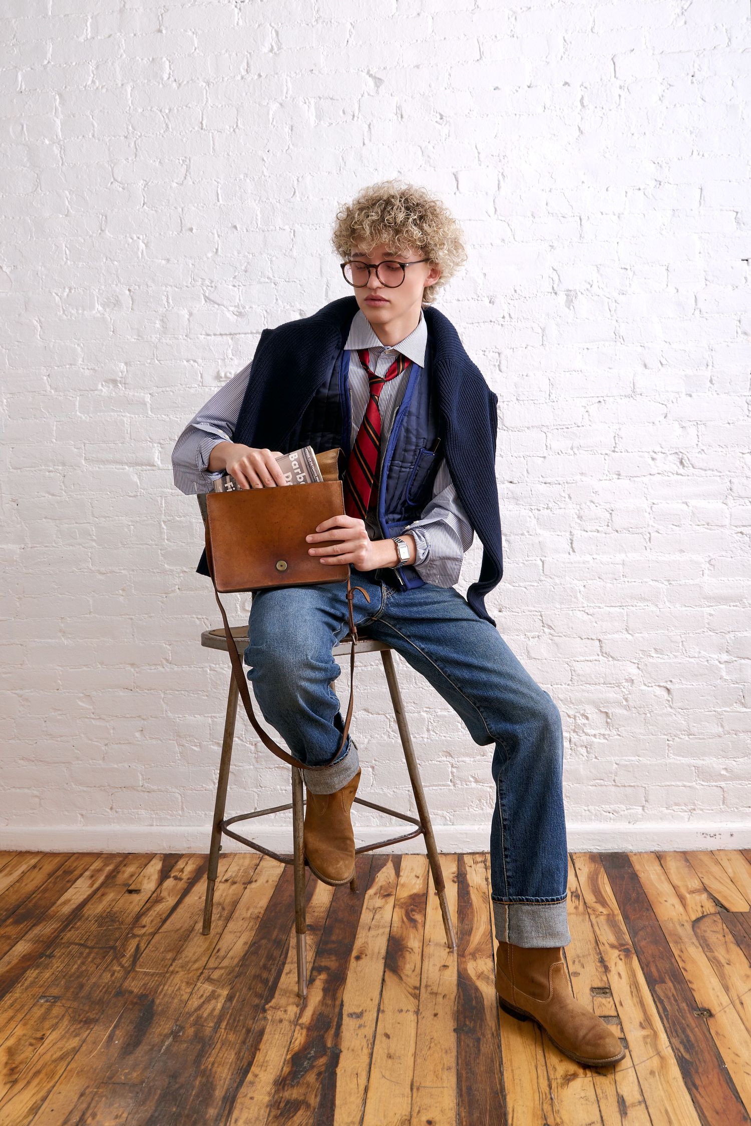 Young man with curly hair and glasses sitting on stool holding leather clutch, wearing jeans, tie, and vest.
