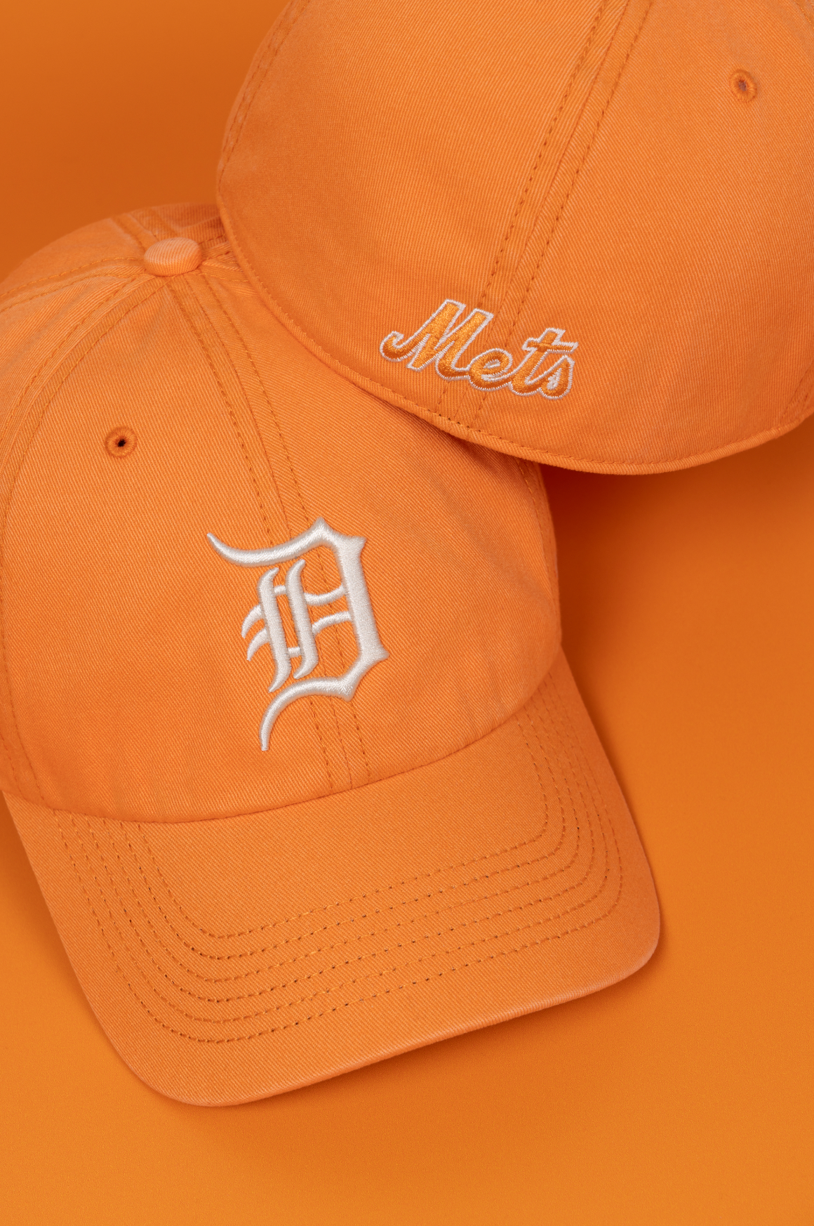 Collection of orange baseball caps with different team logos including Detroit Tigers and New York Mets displayed on white surface.
