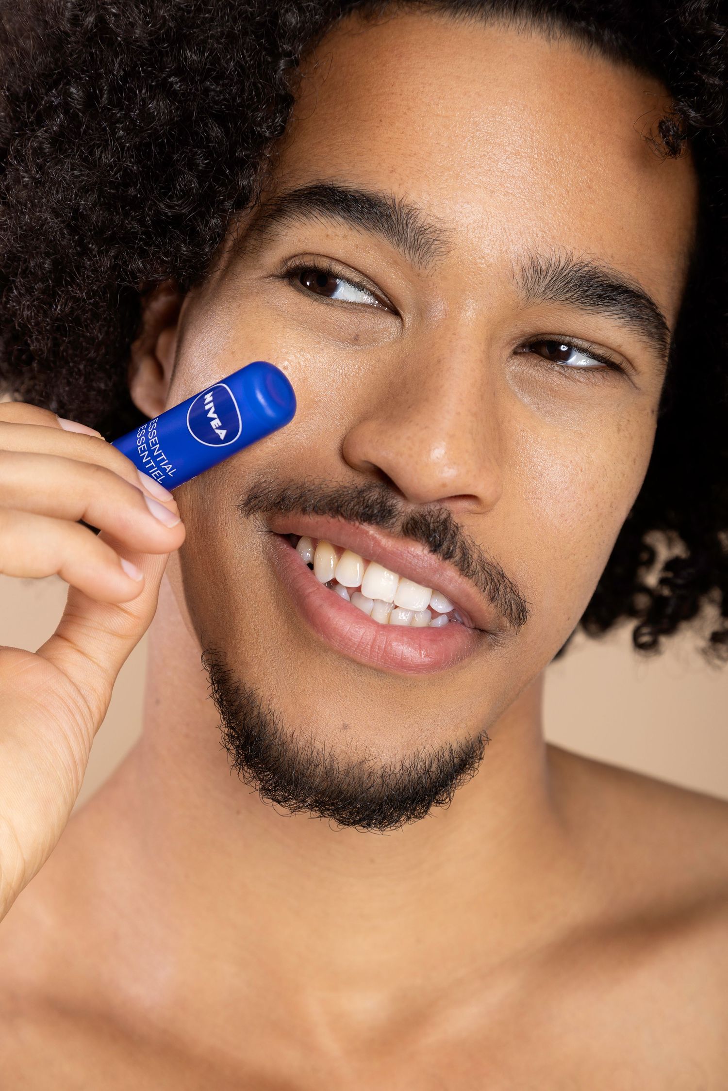 Person with curly hair and facial hair holding a blue container of skin care product against beige background.