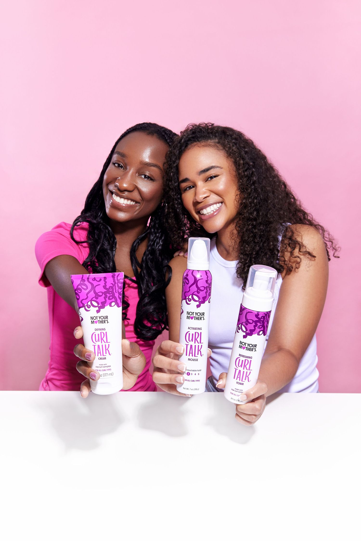 Two women pose with white skincare product bottles against a pink background in a fun promotional photoshoot.