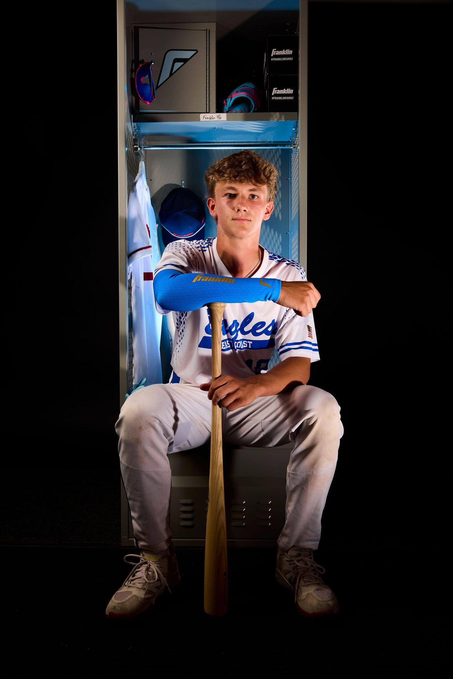 Baseball player in white uniform sits in dark locker room with dramatic lighting and baseball bat.