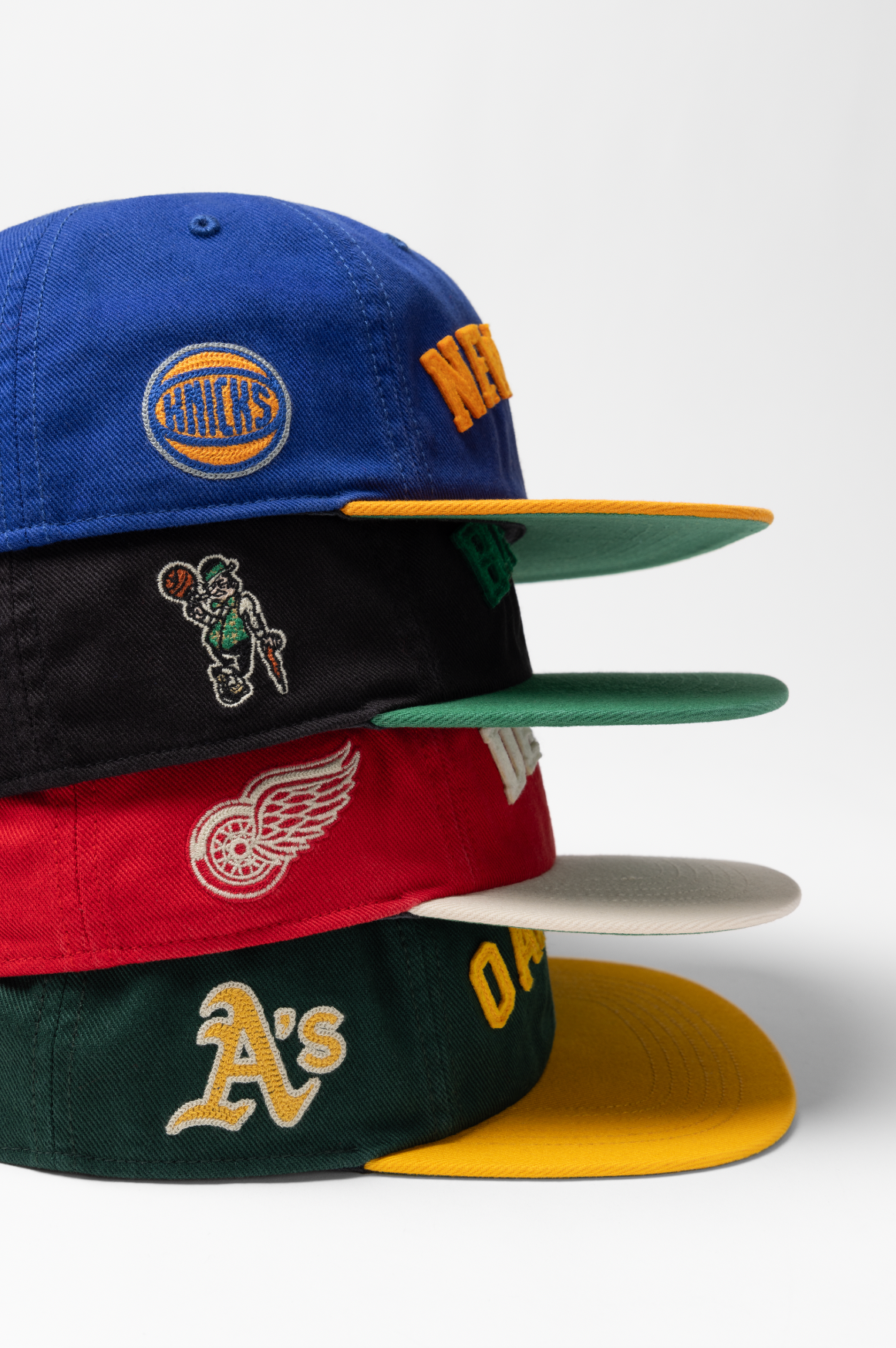 A stack of professional sports team baseball caps featuring the Knicks, Celtics, Red Wings and Athletics logos.