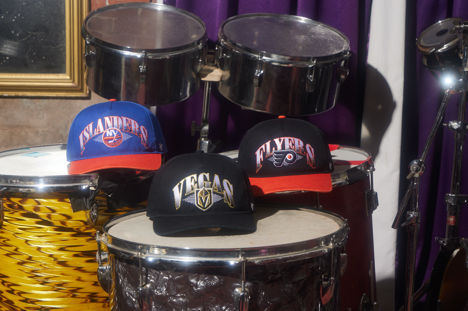 Sports team baseball caps displayed on top of black drums in a music equipment setup.