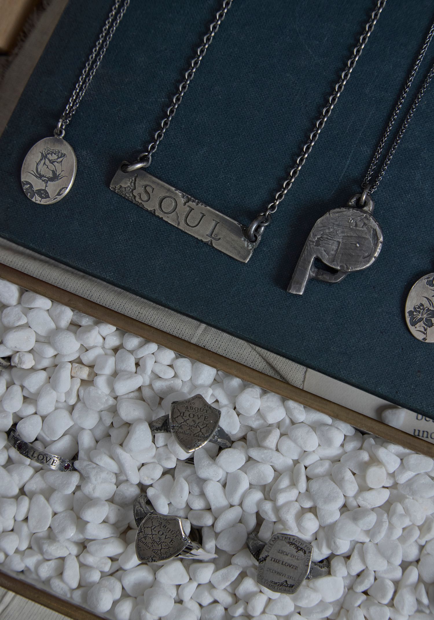 Silver necklaces with coin and bar pendants, including a SOUL bar necklace, displayed with small silver rings on white pebbles.