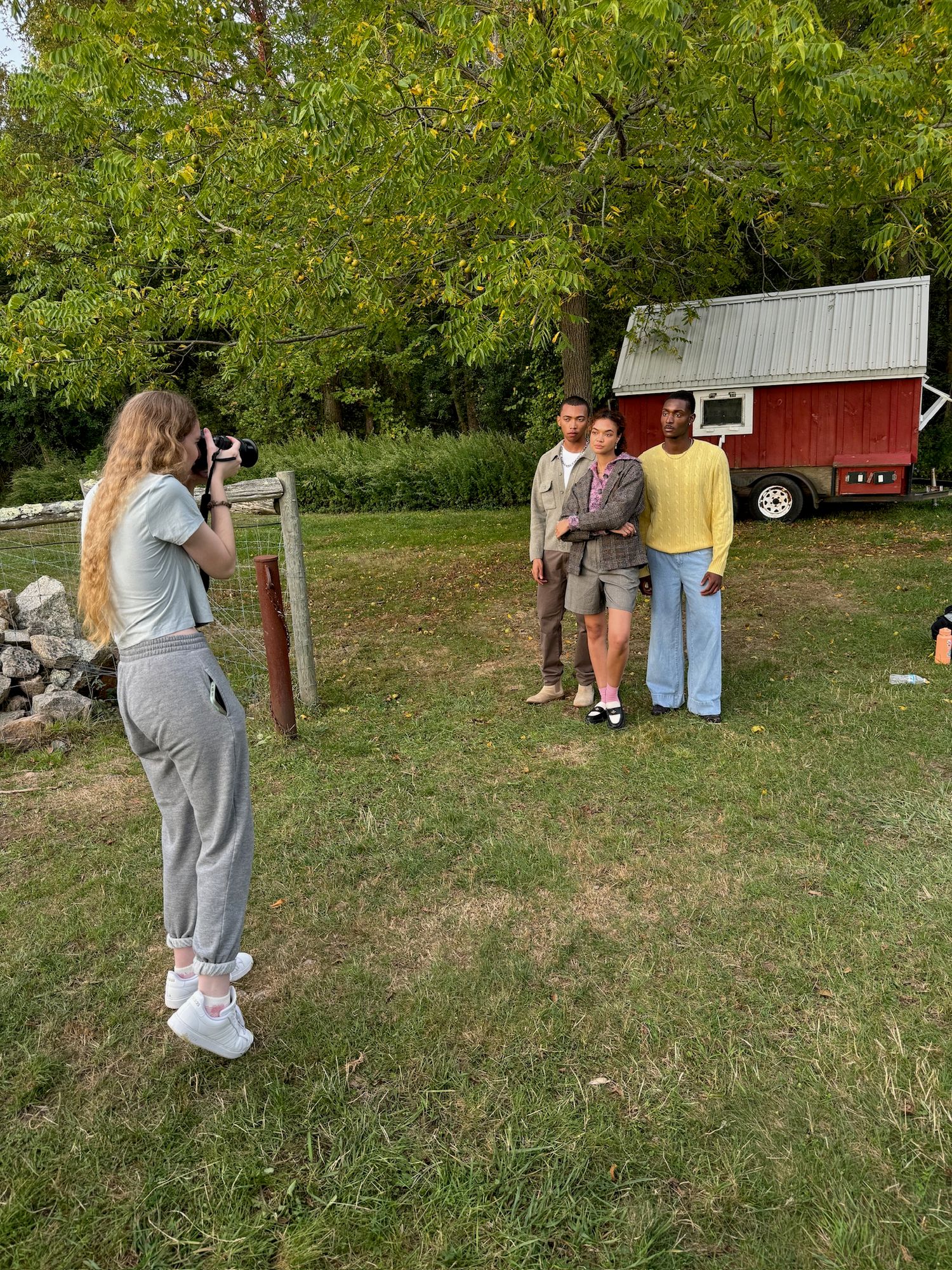 A photographer takes pictures of people posing in front of a red barn on a grassy lawn with autumn trees in the background.
