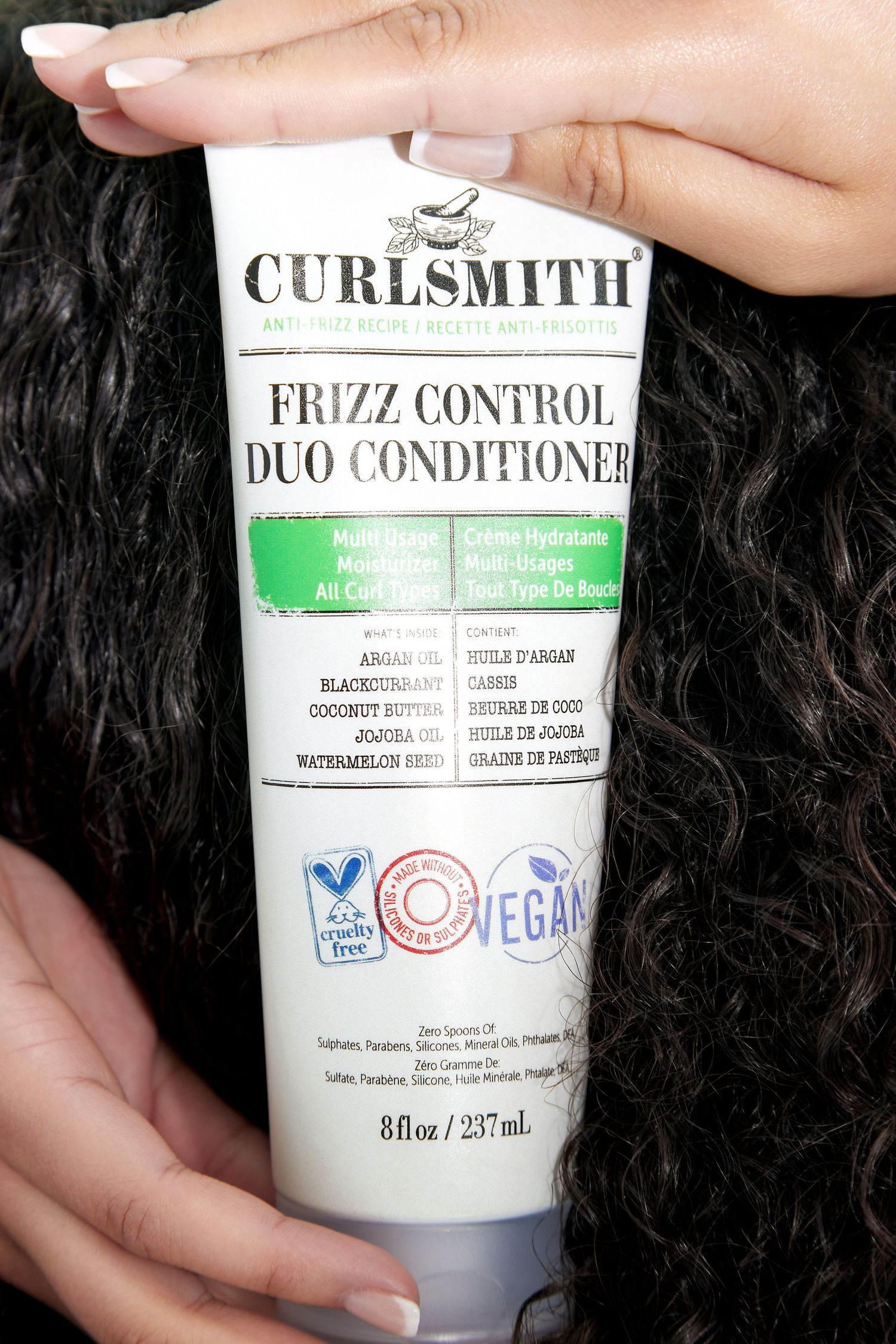 Hands holding and displaying various hair care products including Curlsmith conditioner bottles against dark curly hair background.