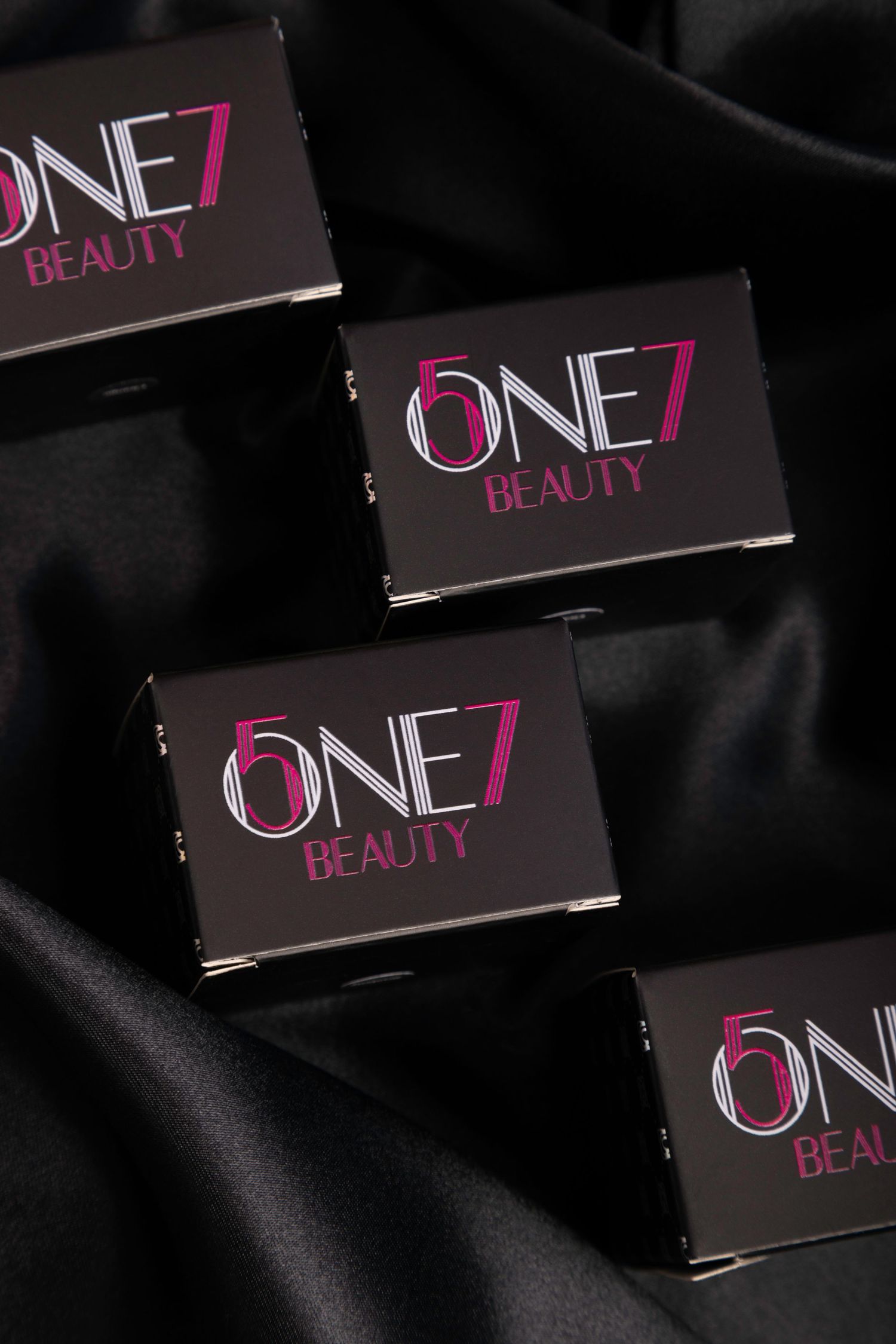 Black and pink ONE7 Beauty branded makeup packaging displayed against dark fabric background.
