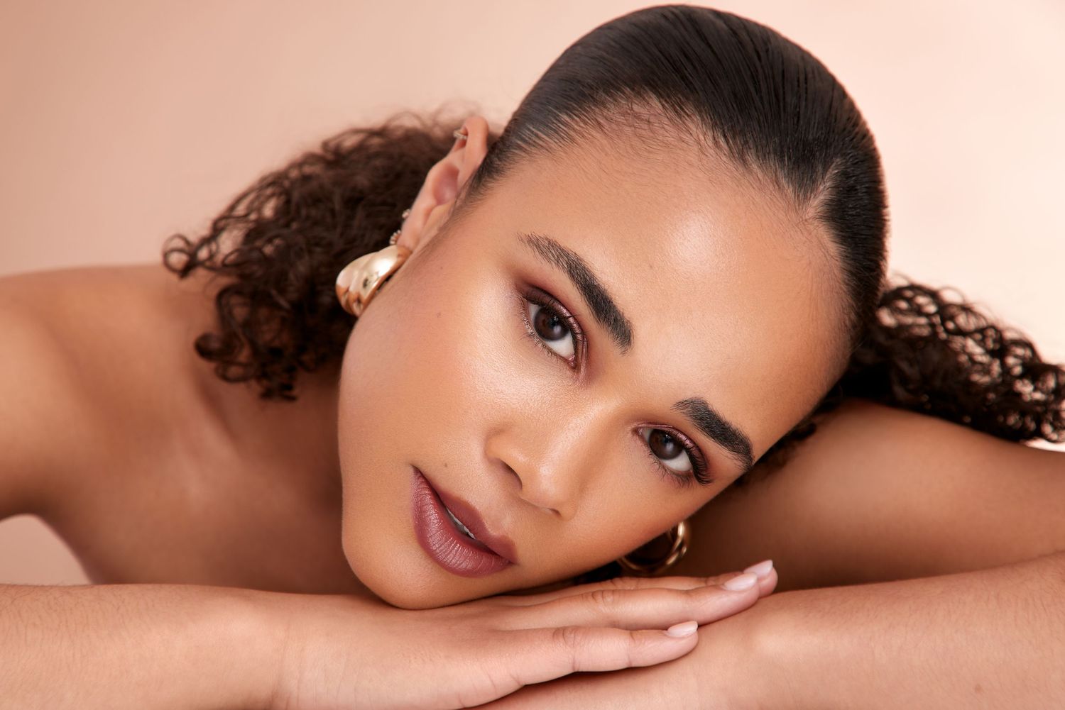 Beauty portrait with natural curly hair and glowing skin resting head on hands against pink background.