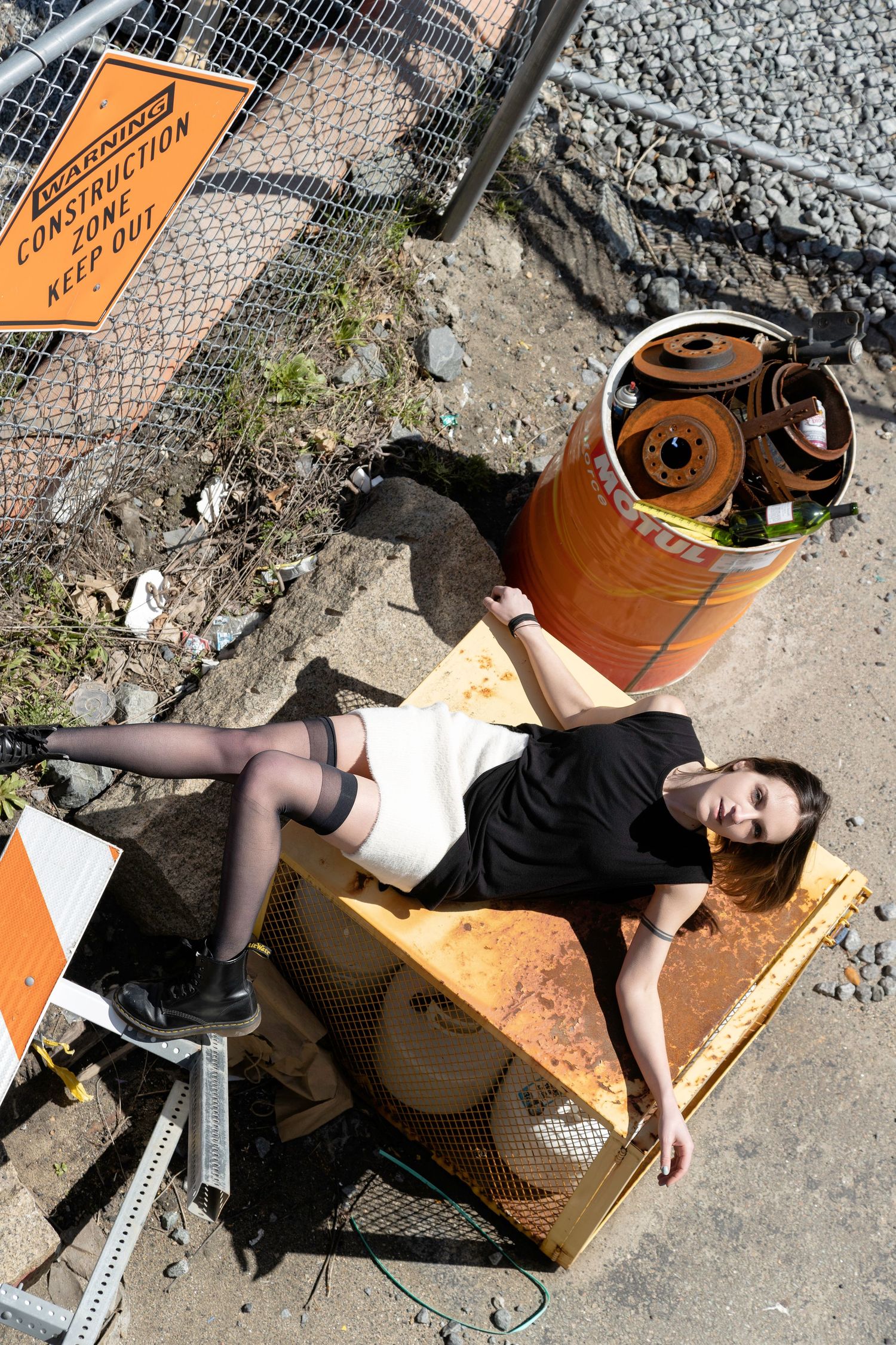 Person in black clothing lying on ground next to orange cardboard boxes and chain link fence in urban setting.