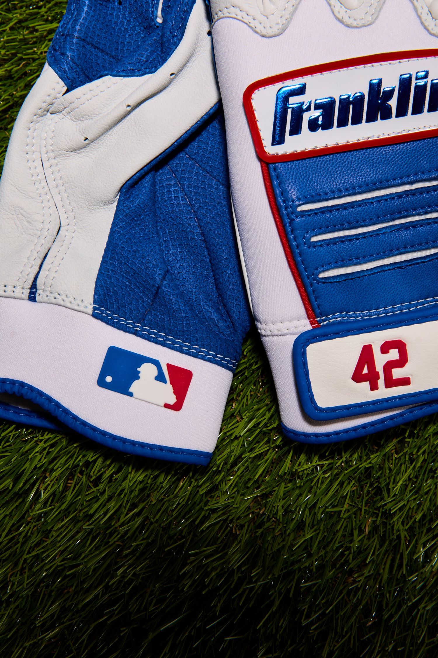 Close-up view of white and blue Franklin baseball batting gloves with red accents laying on artificial turf.