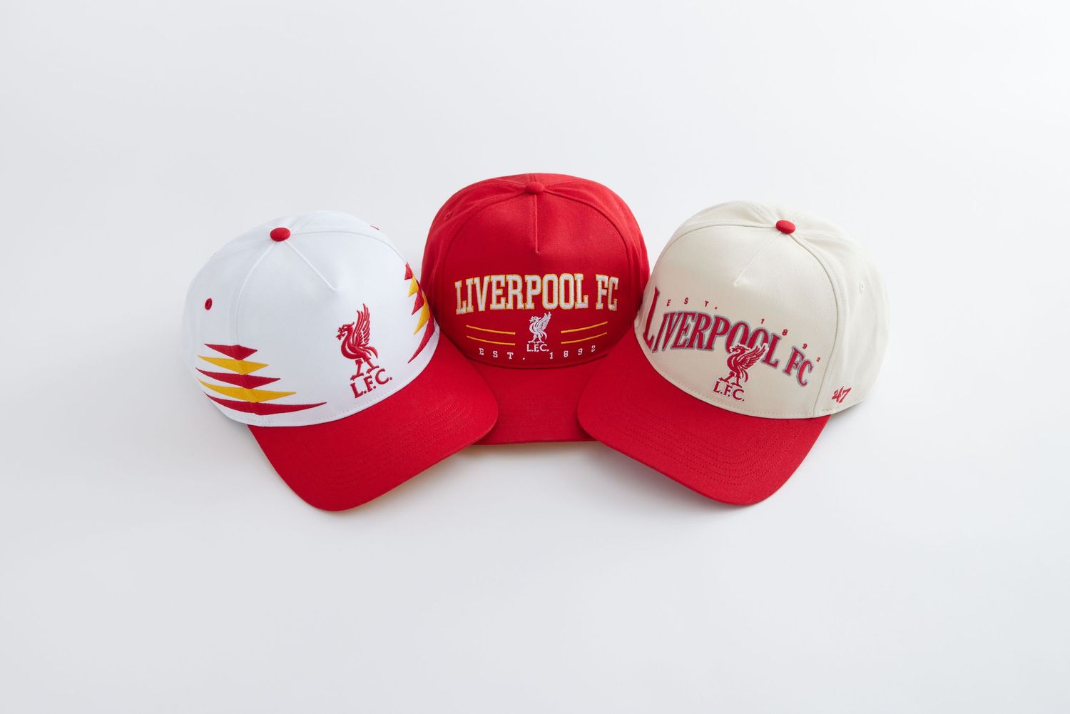 Collection of red and white baseball caps featuring Liverpool FC and other sports team logos displayed on white background.