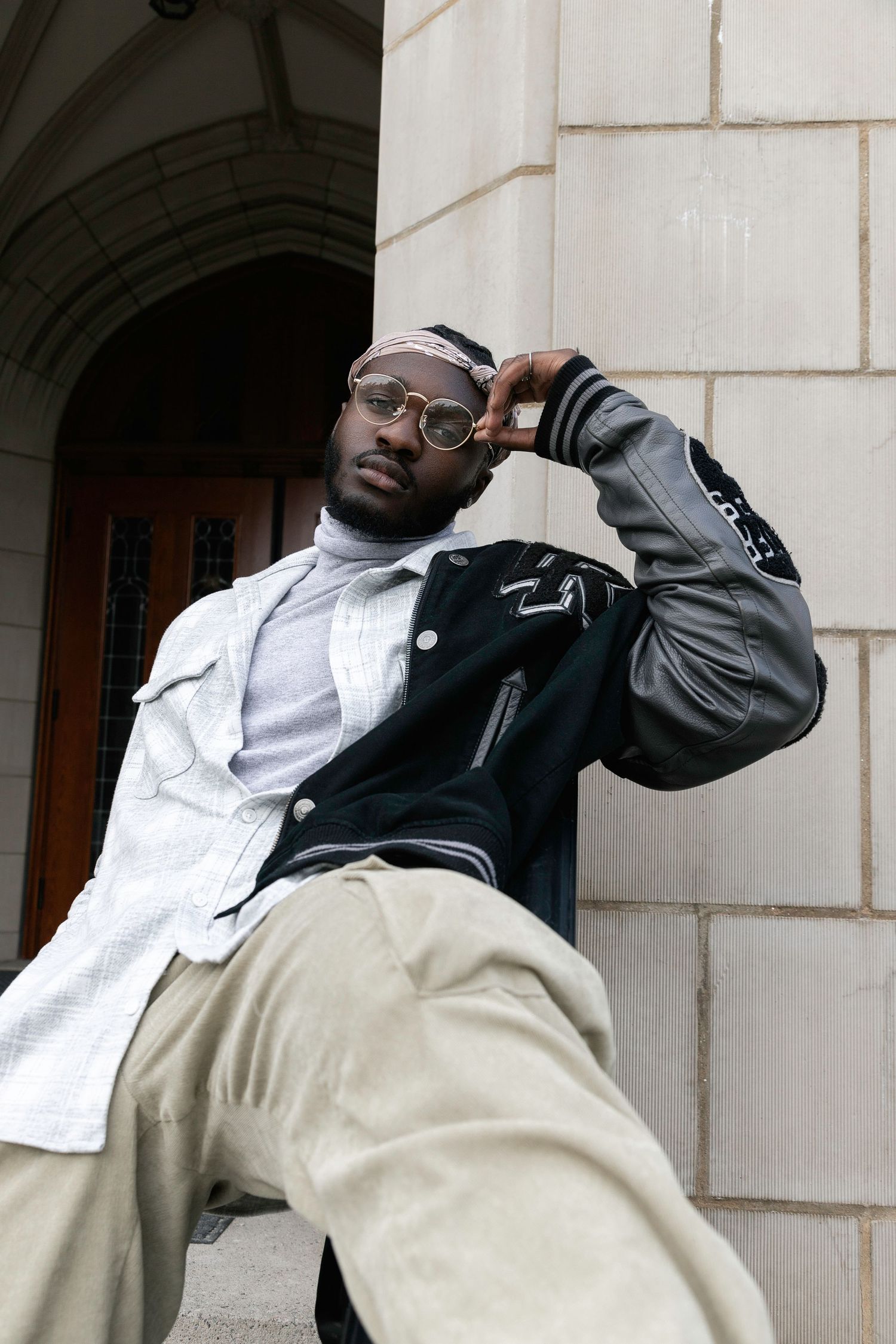 Person in black jacket and white shirt poses against stone wall in architectural setting.