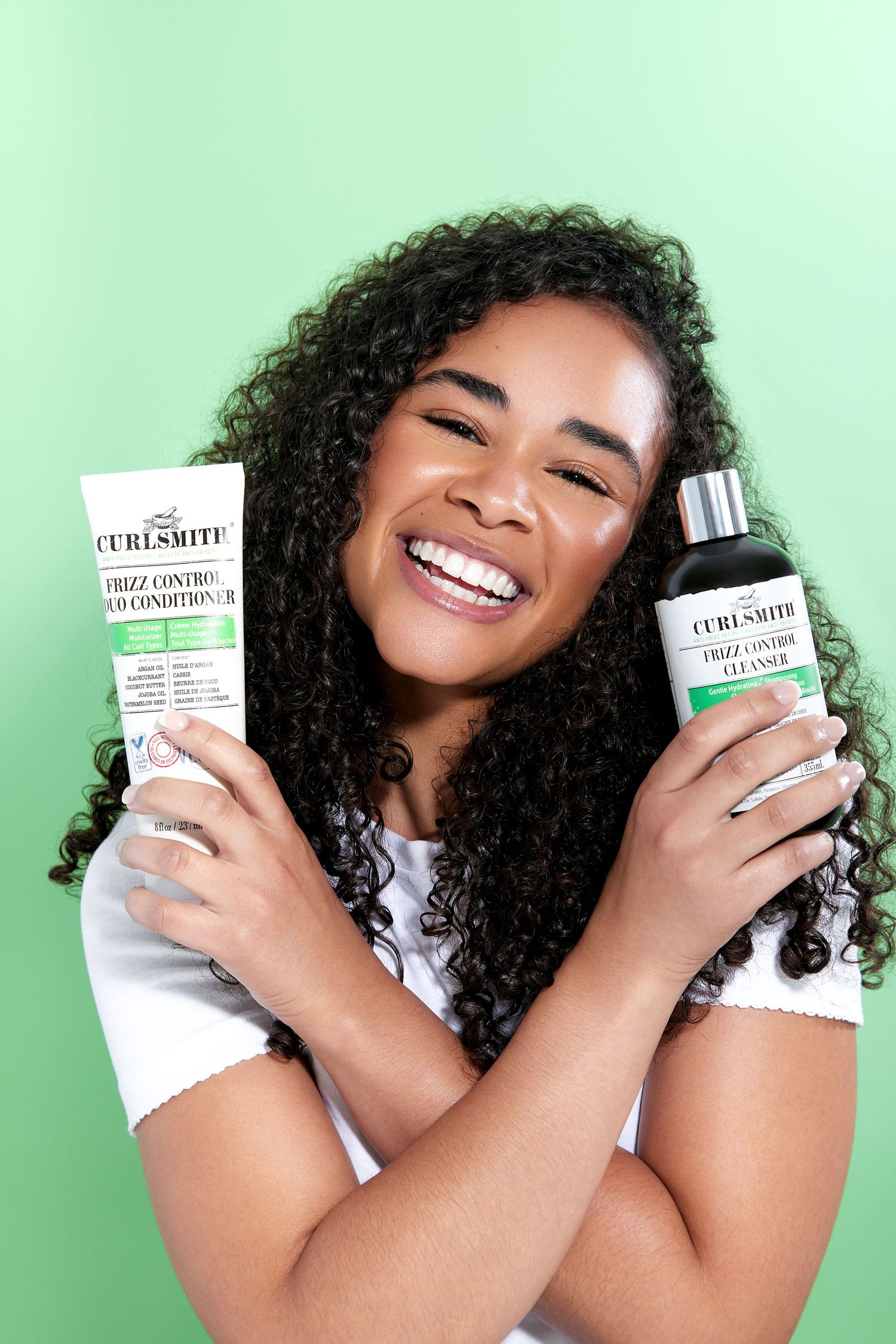 A smiling woman with curly hair holds up skincare product bottles against a mint green background.