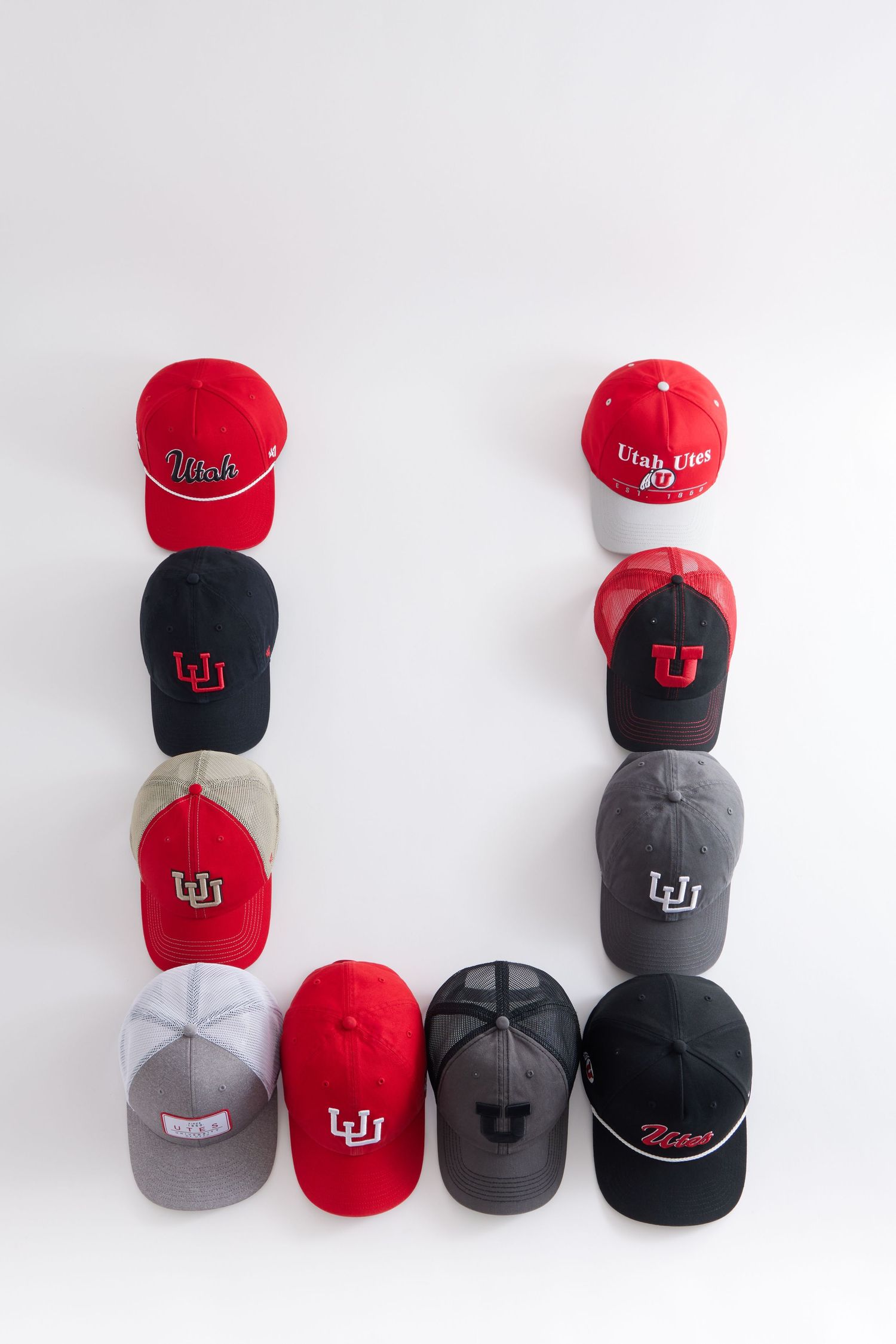 Multiple University of Utah baseball caps in red, black, and gray colors arranged in different stacks and groupings.