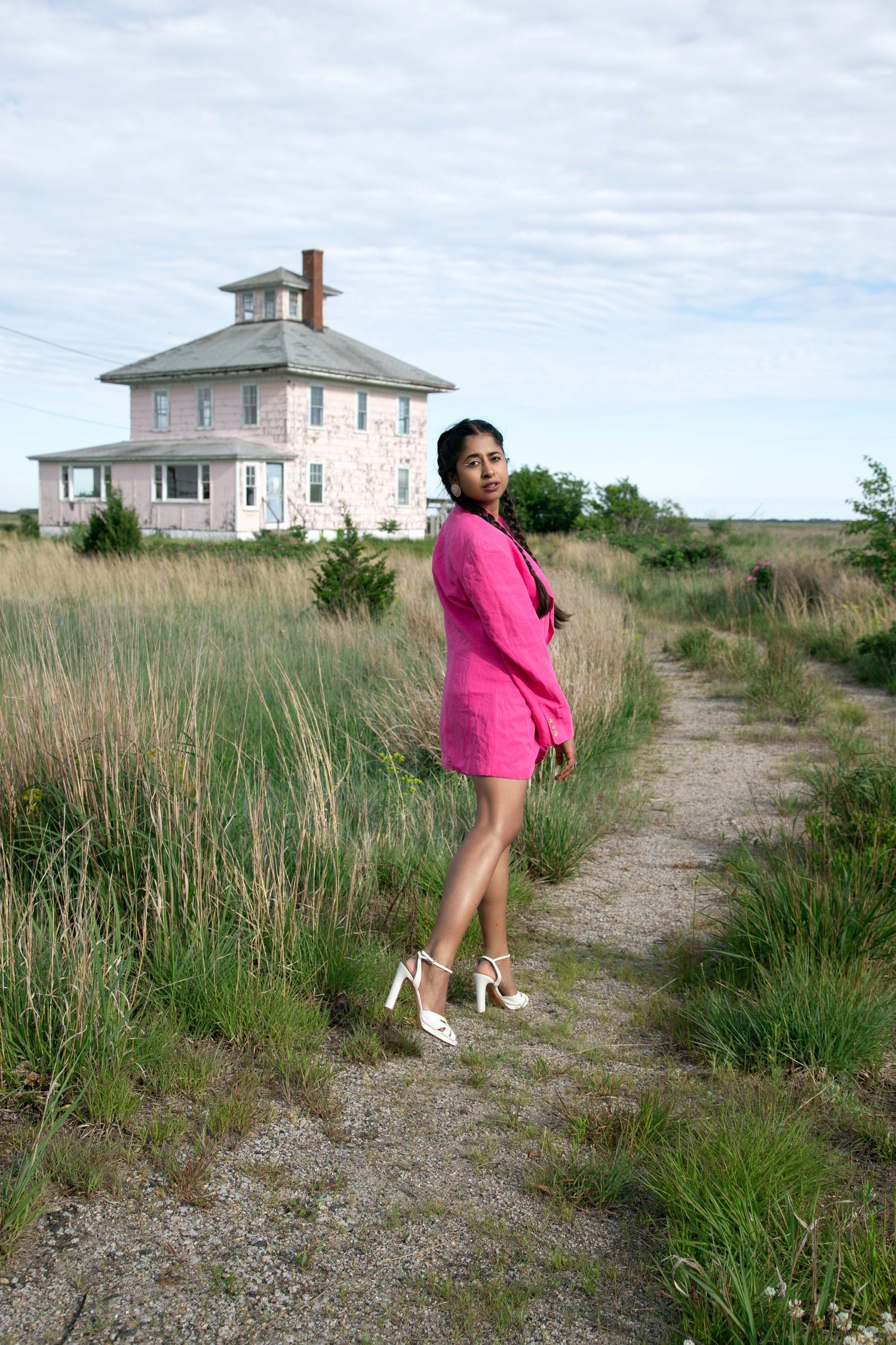 A woman in a bright pink dress poses playfully along a grassy path leading to a coastal house on a cloudy day.