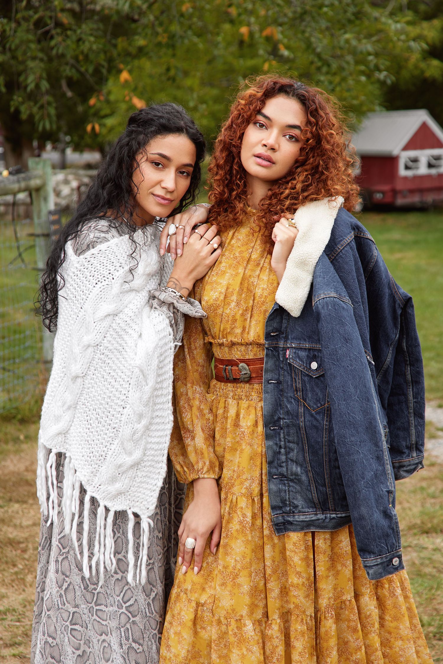 Two people wearing contrasting outfits of a white crochet shawl and yellow floral dress with denim jacket pose together outdoors.
