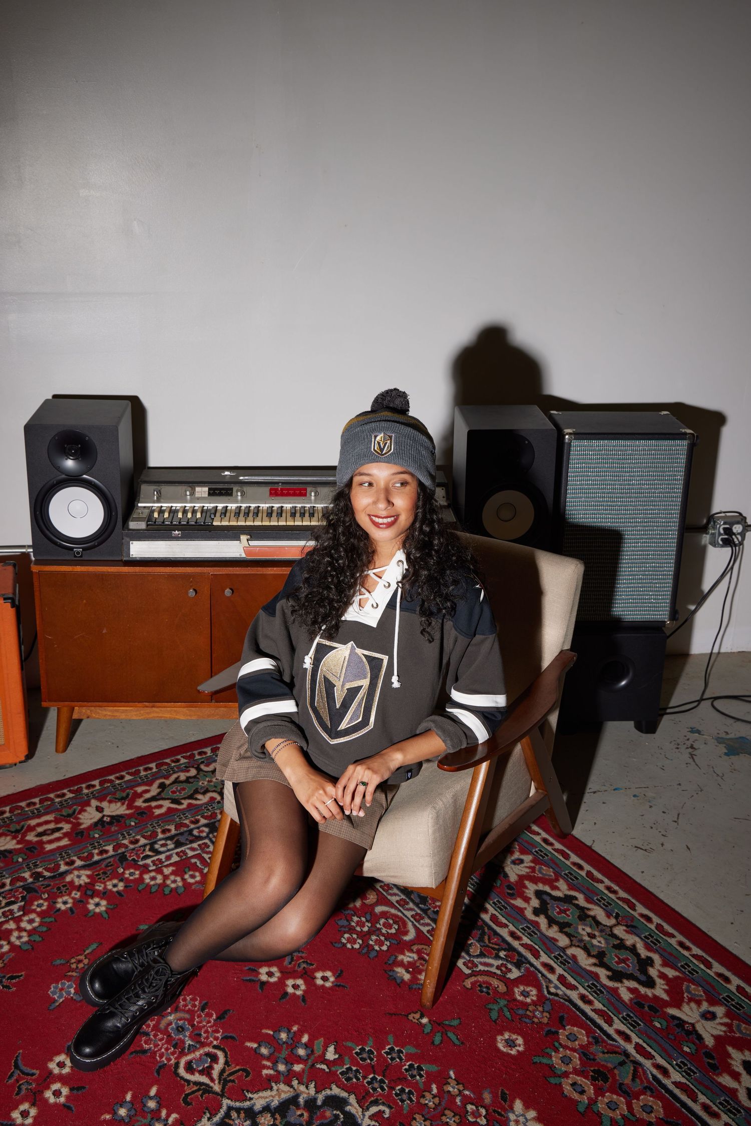 Woman wearing black baseball cap and Vegas Golden Knights jersey lounges on red patterned carpet in room with audio equipment.
