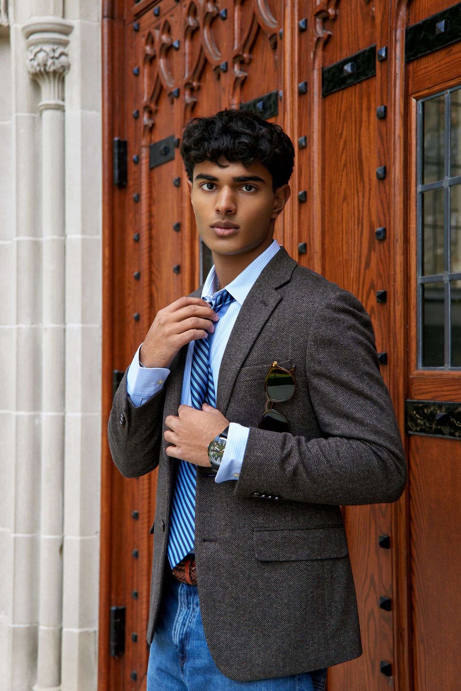 A person in a grey suit jacket and blue striped tie poses against ornate wooden doors with decorative metal hardware.