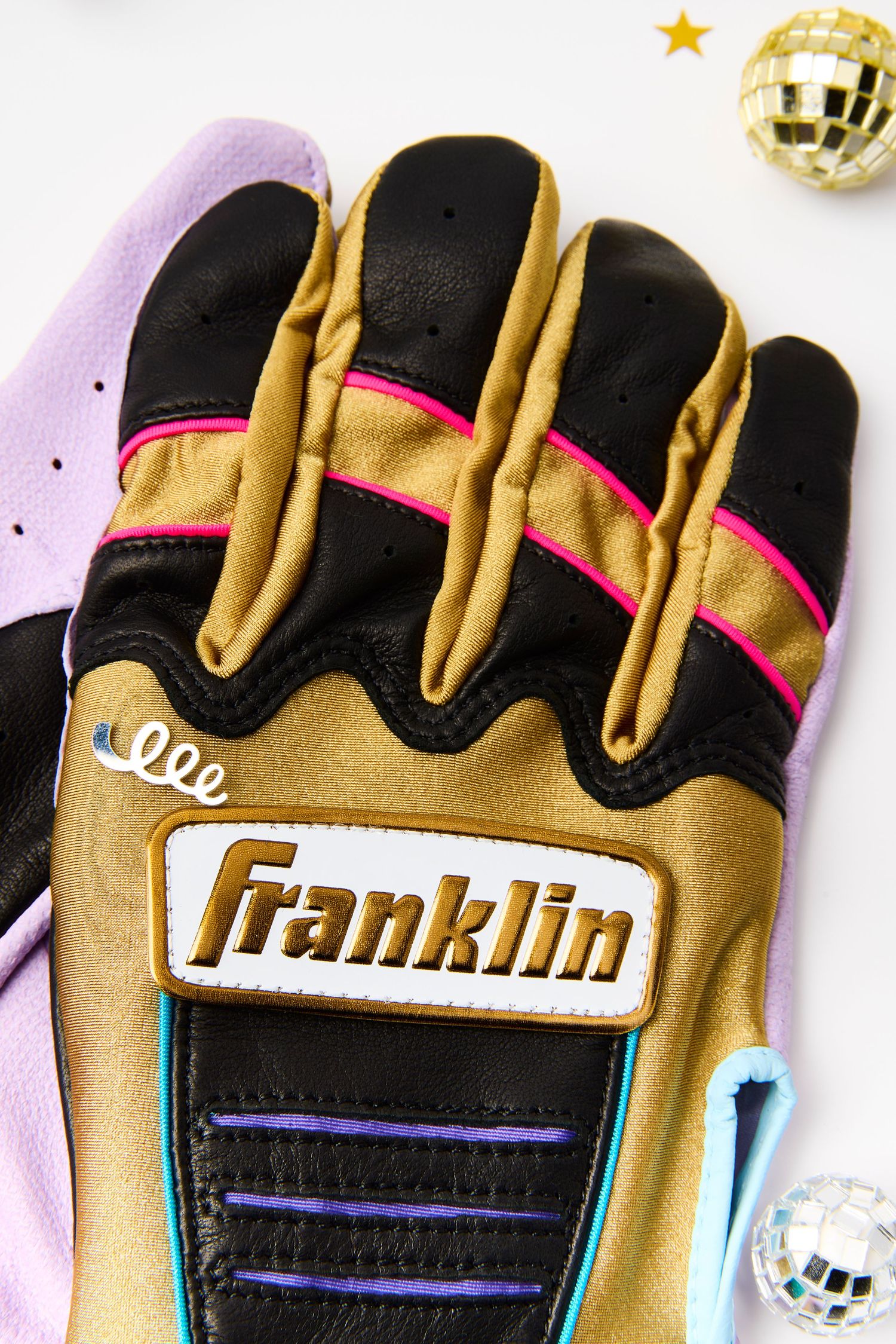 A gold and black Franklin sports glove with pink accents and purple stripes shown against a white background with disco balls.