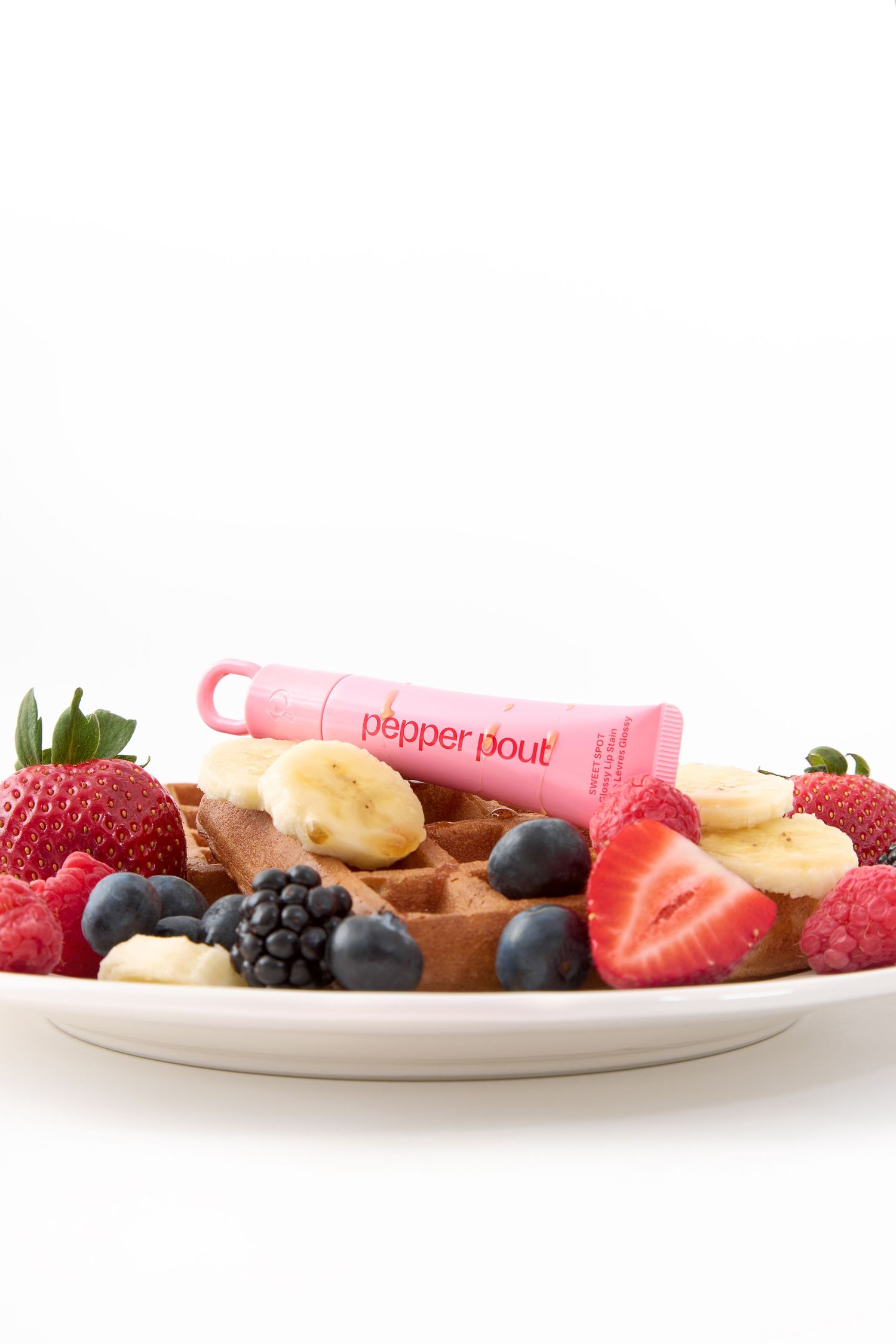 Pepper Pout lip gloss tube displayed on a waffle plate with fresh strawberries, blueberries, raspberries, and banana slices.