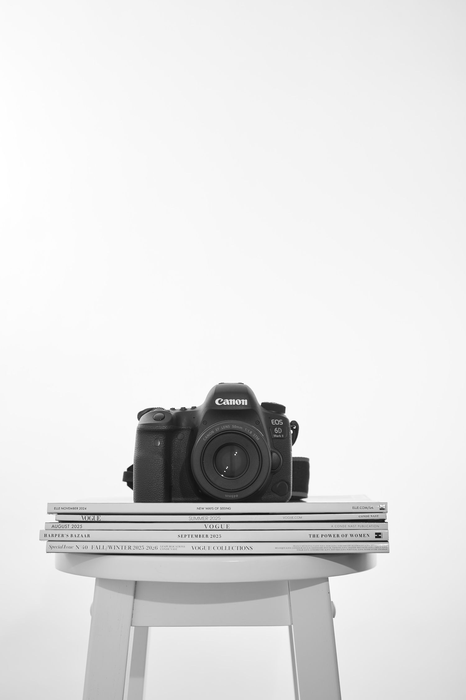 A Canon DSLR camera sits atop white magazines and books on a minimalist white table in a clean monochromatic setting.