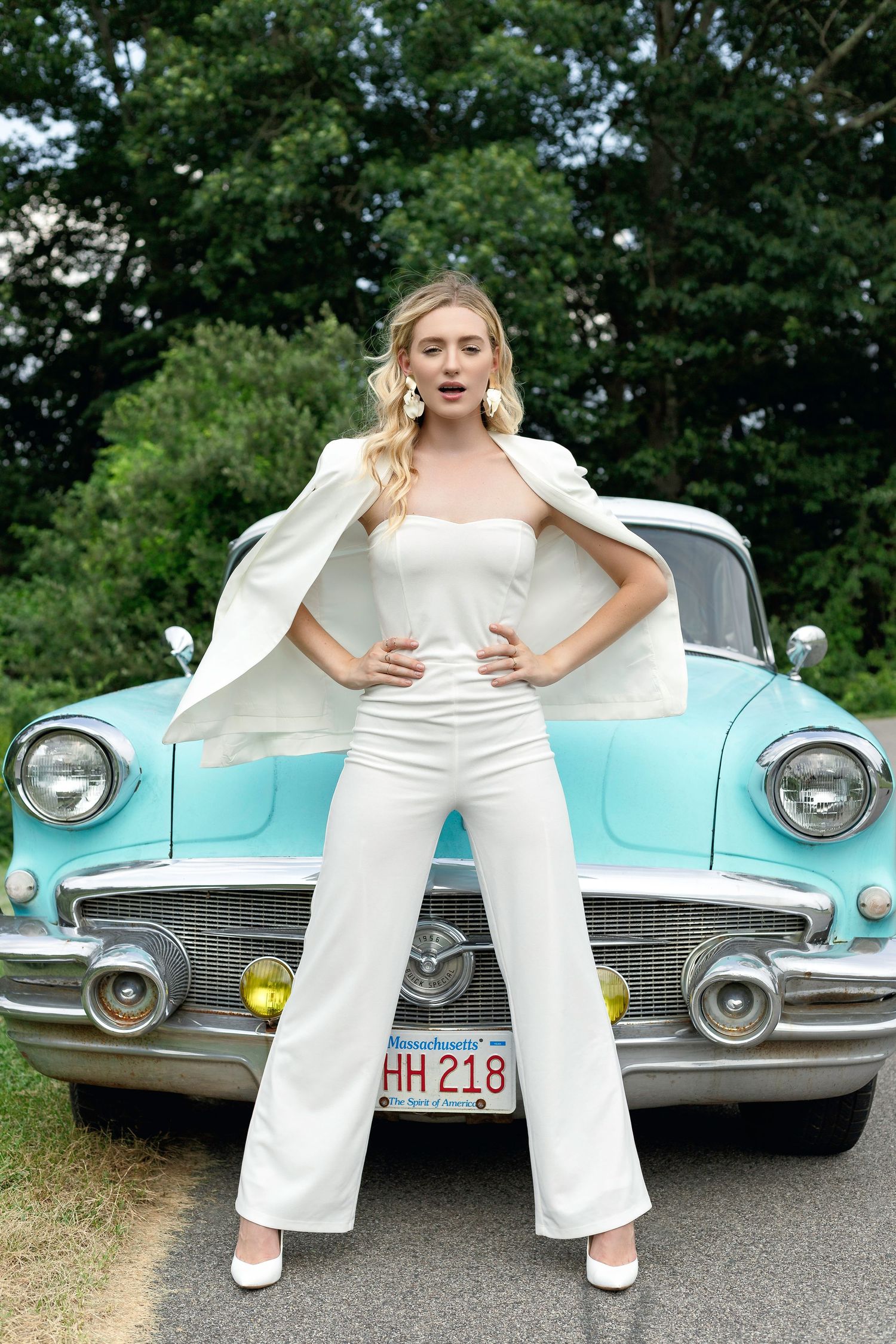 A person in white attire poses playfully in front of a vintage turquoise classic car in an outdoor setting.