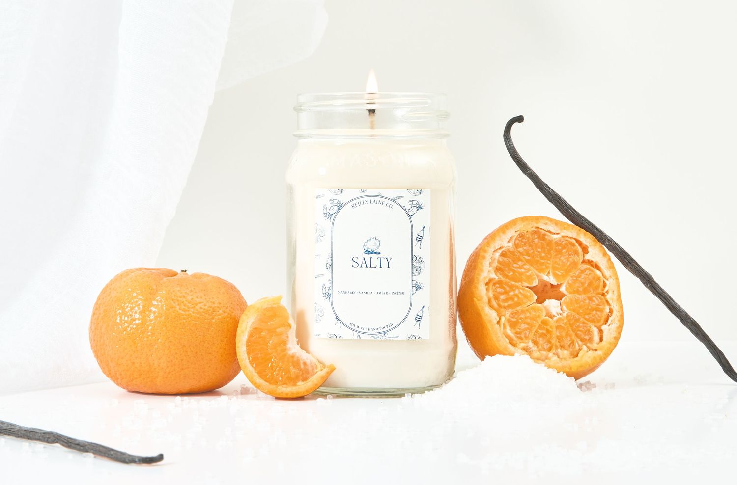 White jar candle with orange vanilla scent, surrounded by fresh tangerines and vanilla beans on white background.