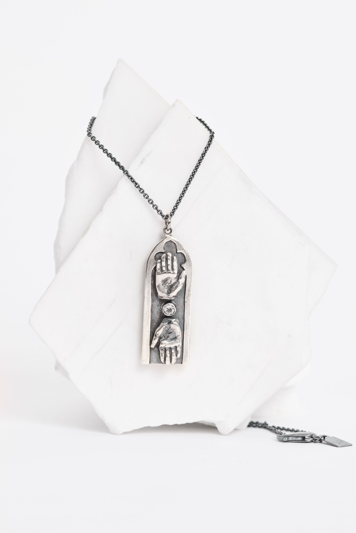 Silver rectangular pendant necklace with hand and eye motifs on dark chain, displayed on white marble.