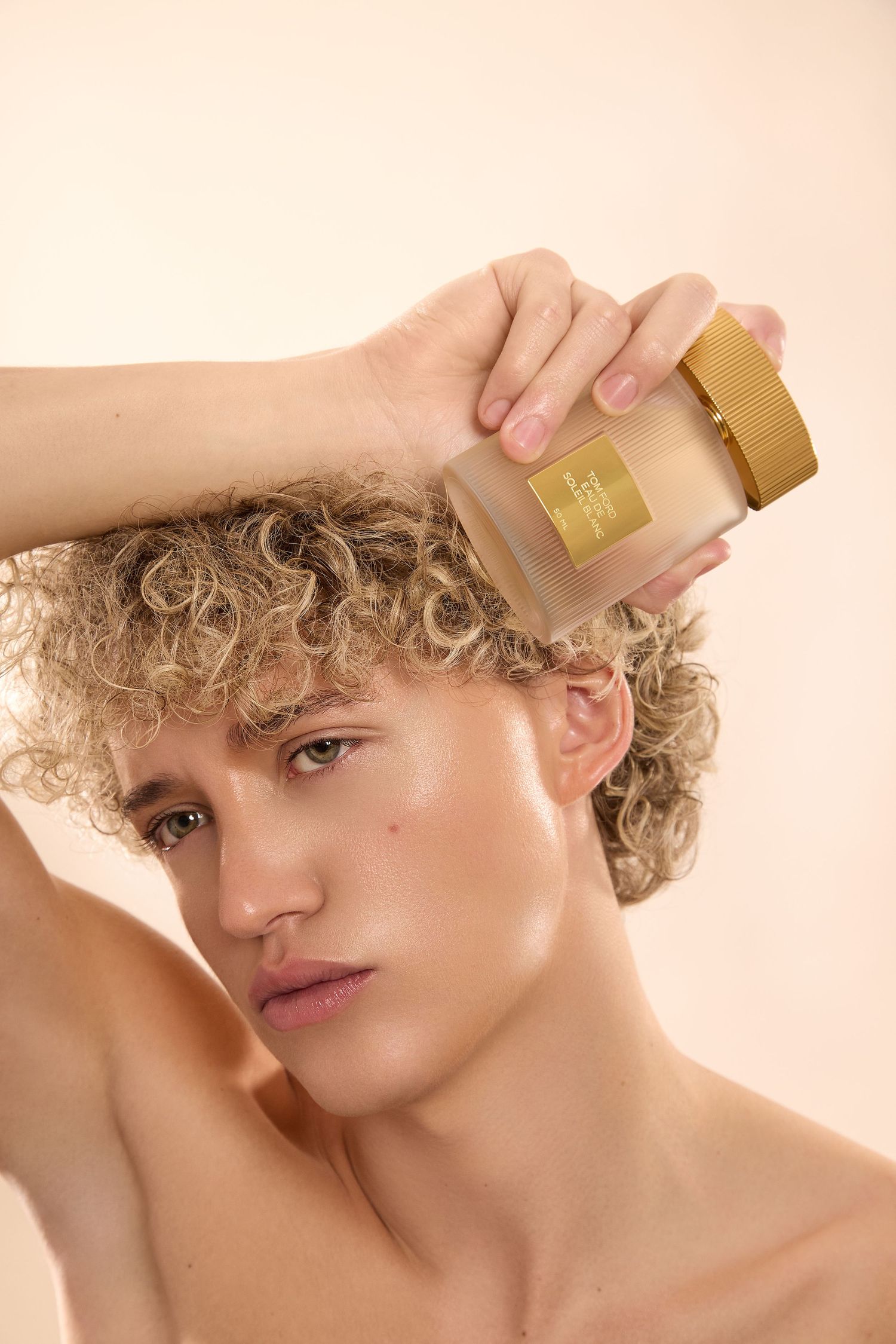 Young man with curly blonde hair holding a gold luxury perfume bottle against his head on a beige background.