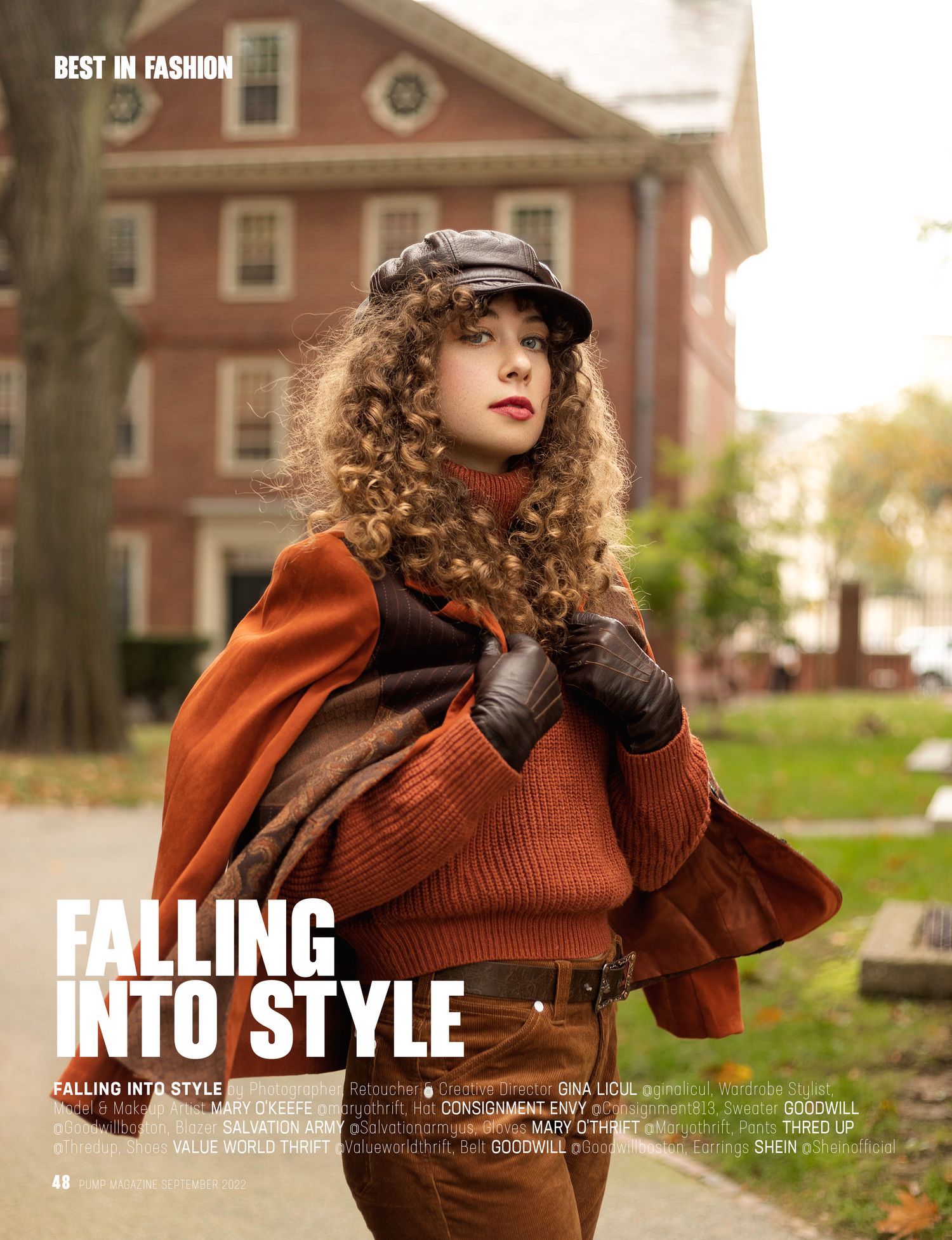 Fashion model in rustic orange sweater and leather gloves poses outdoors against brick building at sunset.