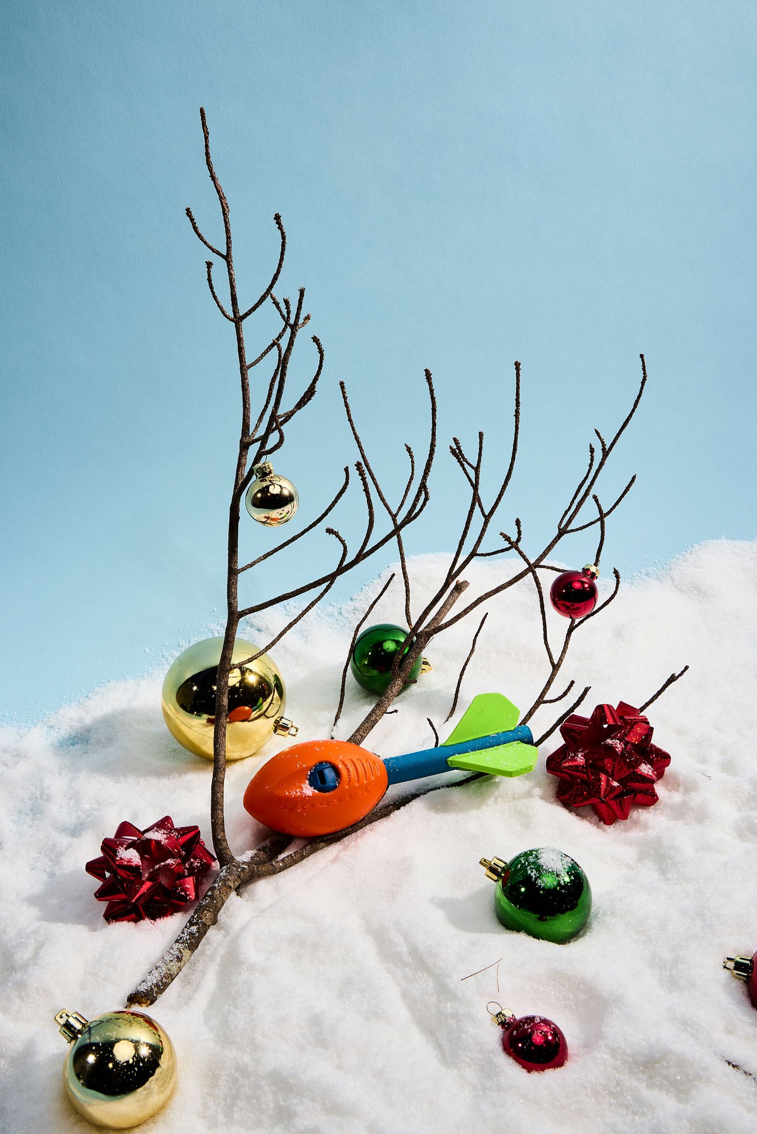 A sequence of colorful Christmas ornaments and bare branches arranged artistically in snow against a blue sky background.