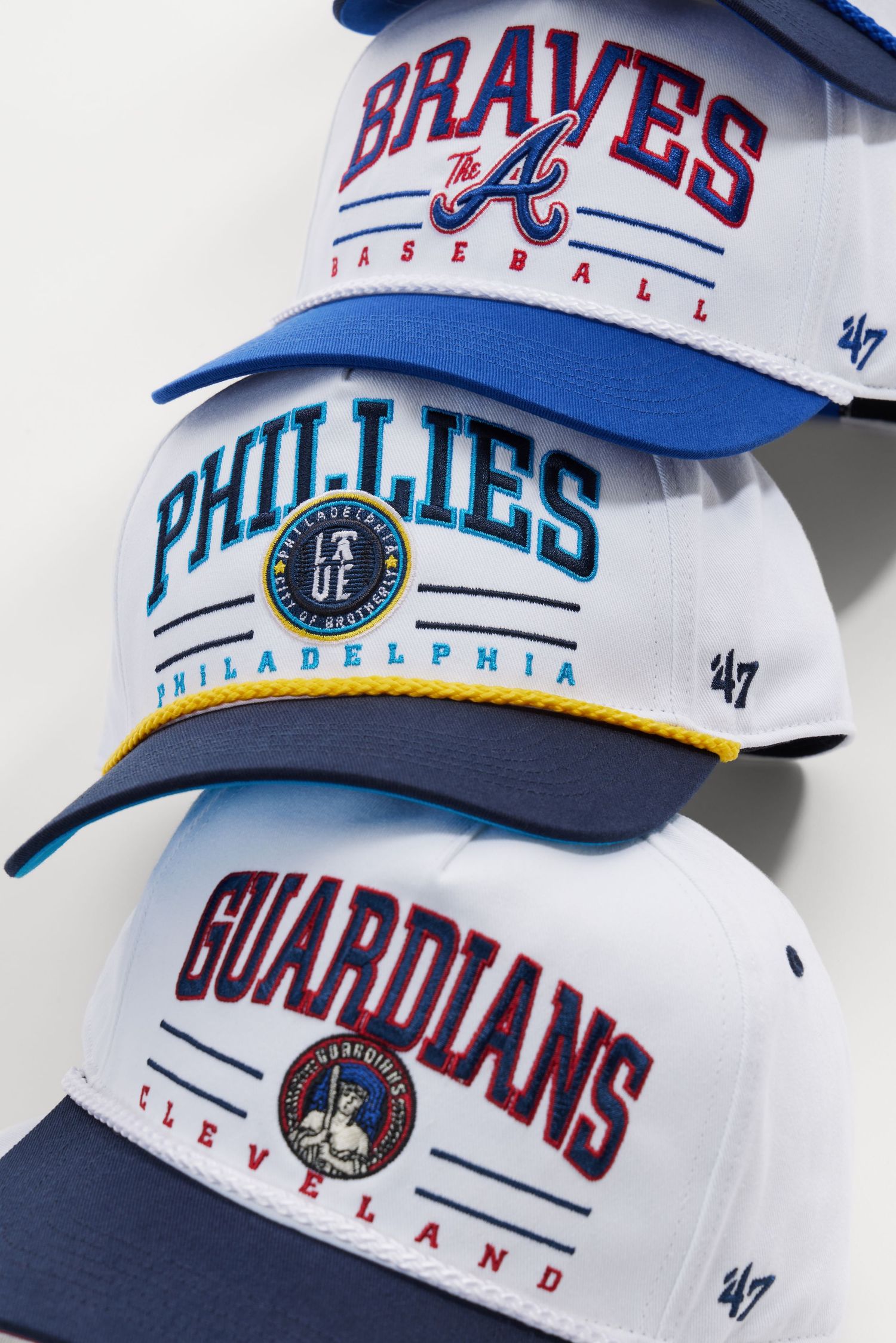 Collection of MLB baseball caps featuring team logos including Braves, Phillies, and Guardians displayed on white surface.