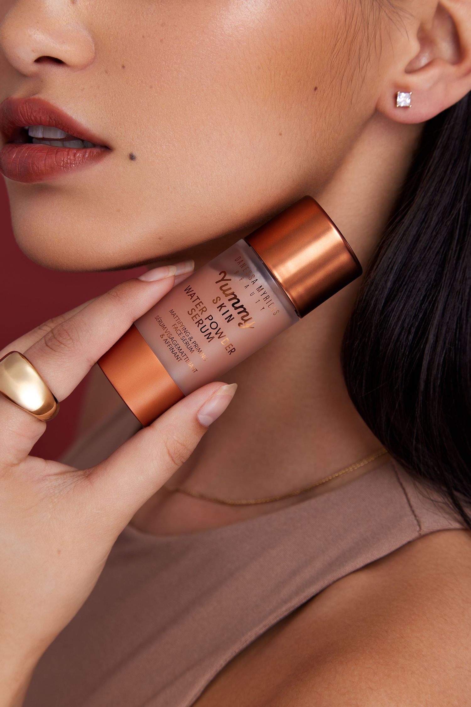 Close-up beauty shot of bronze makeup product bottle held against model's neck with manicured hand.