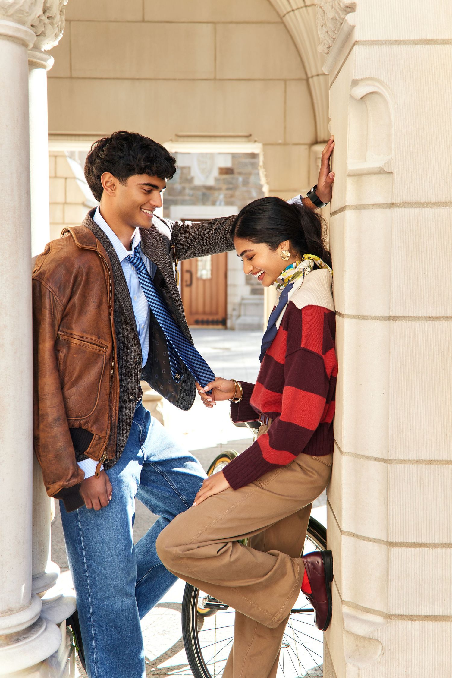 Two students in casual fall attire having a playful moment between stone pillars at a university campus.