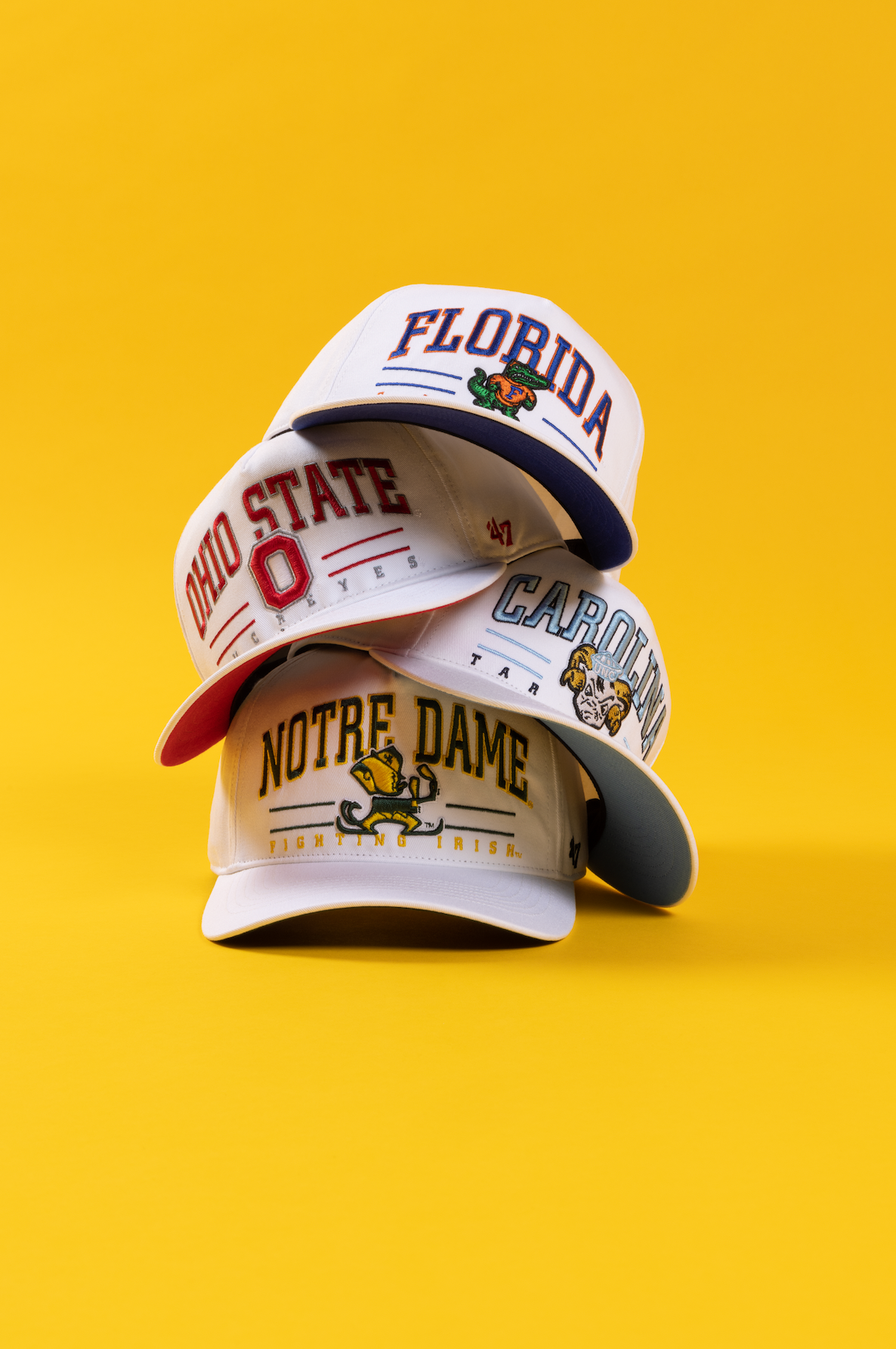 Collection of white Notre Dame baseball caps with team logos displayed on bright yellow background.