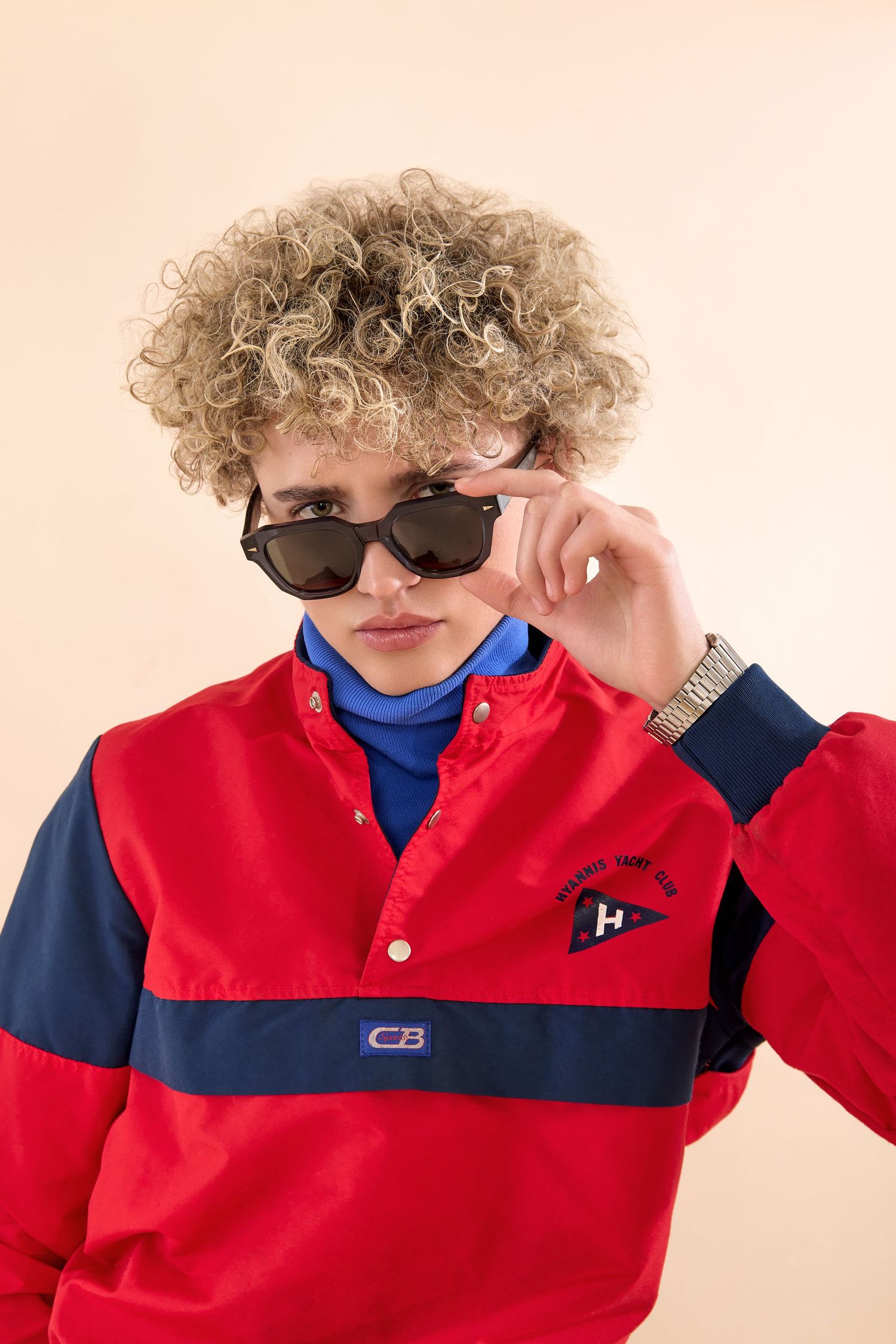 Young man with curly blonde hair wearing red and navy pullover jacket and sunglasses, silver watch on wrist.