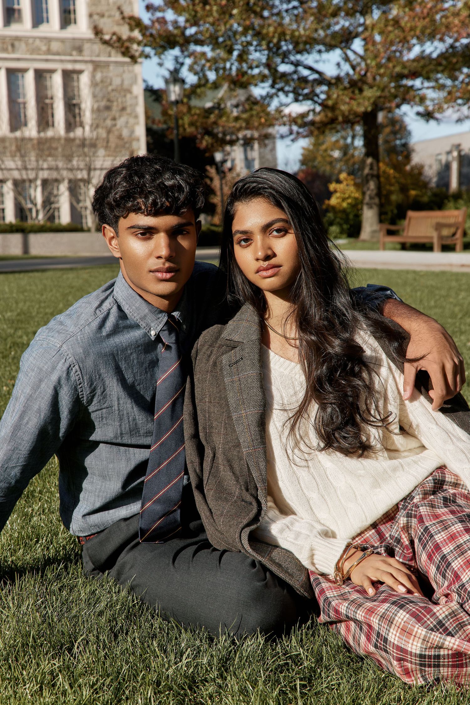 A couple lounges on grass in autumn wearing coordinated outfits of grey suit and plaid skirt with white sweater.