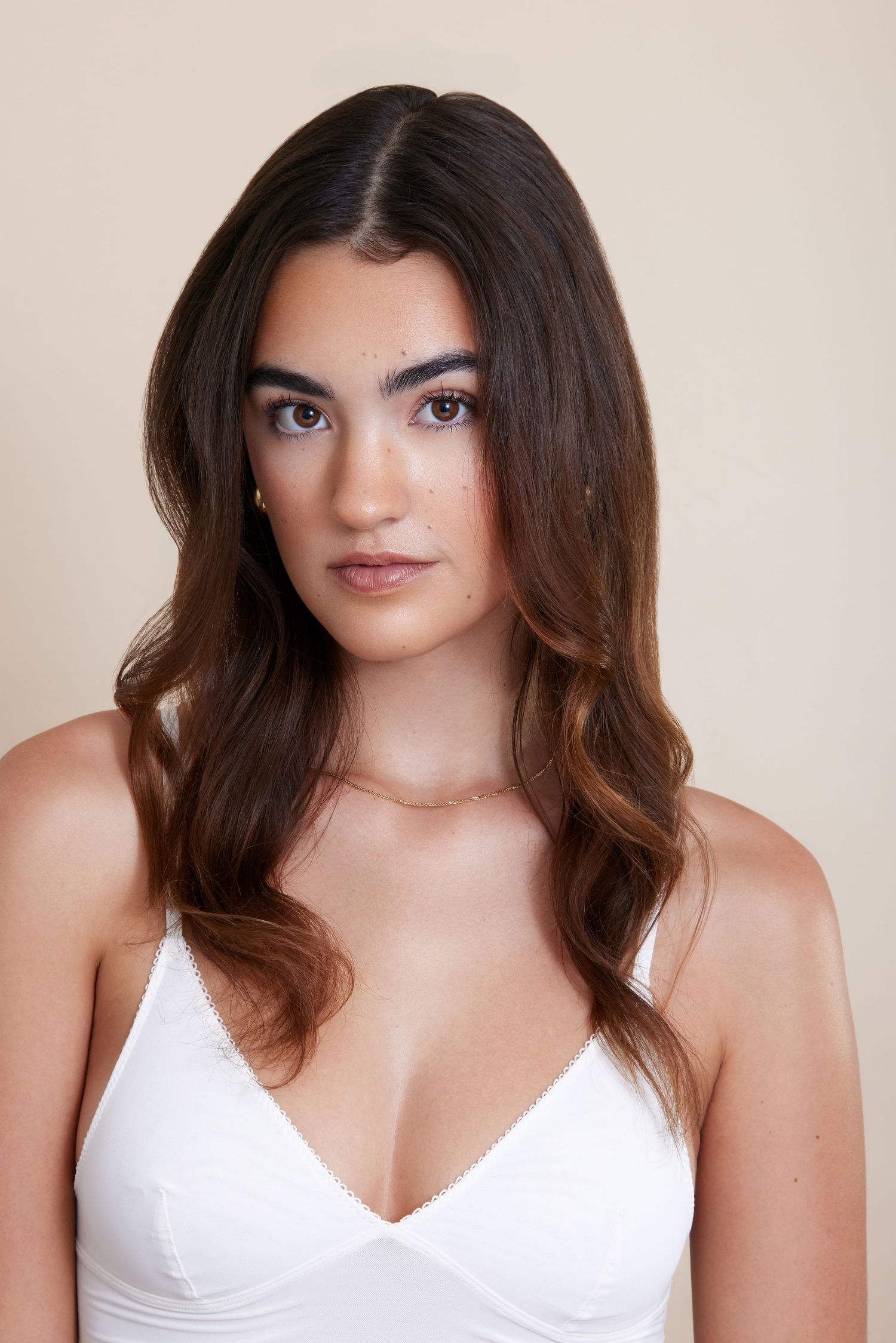 Woman with long dark brown hair wearing white top poses in studio against neutral background.