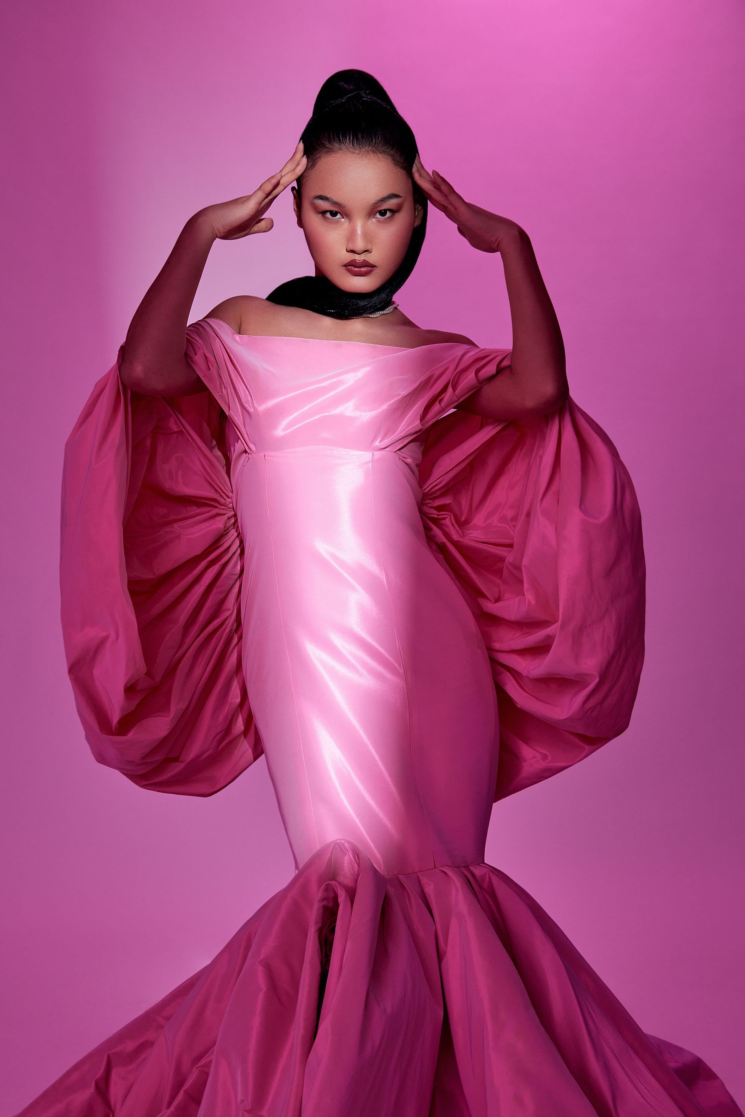 Fashion model poses in dramatic pink mermaid gown with oversized sleeves against magenta backdrop.