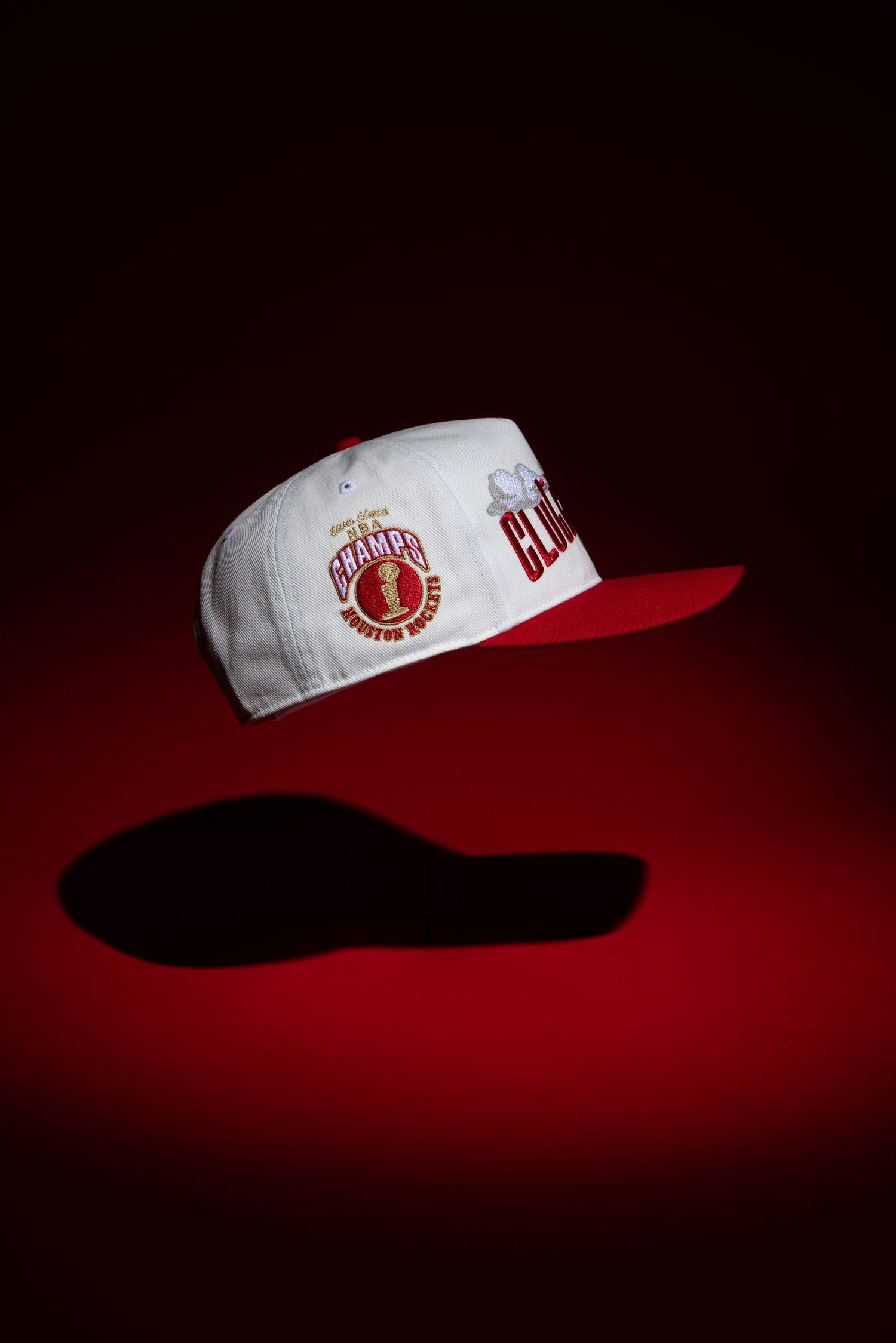 A white and red baseball cap with a logo floating against a dark red background with dramatic shadow beneath.